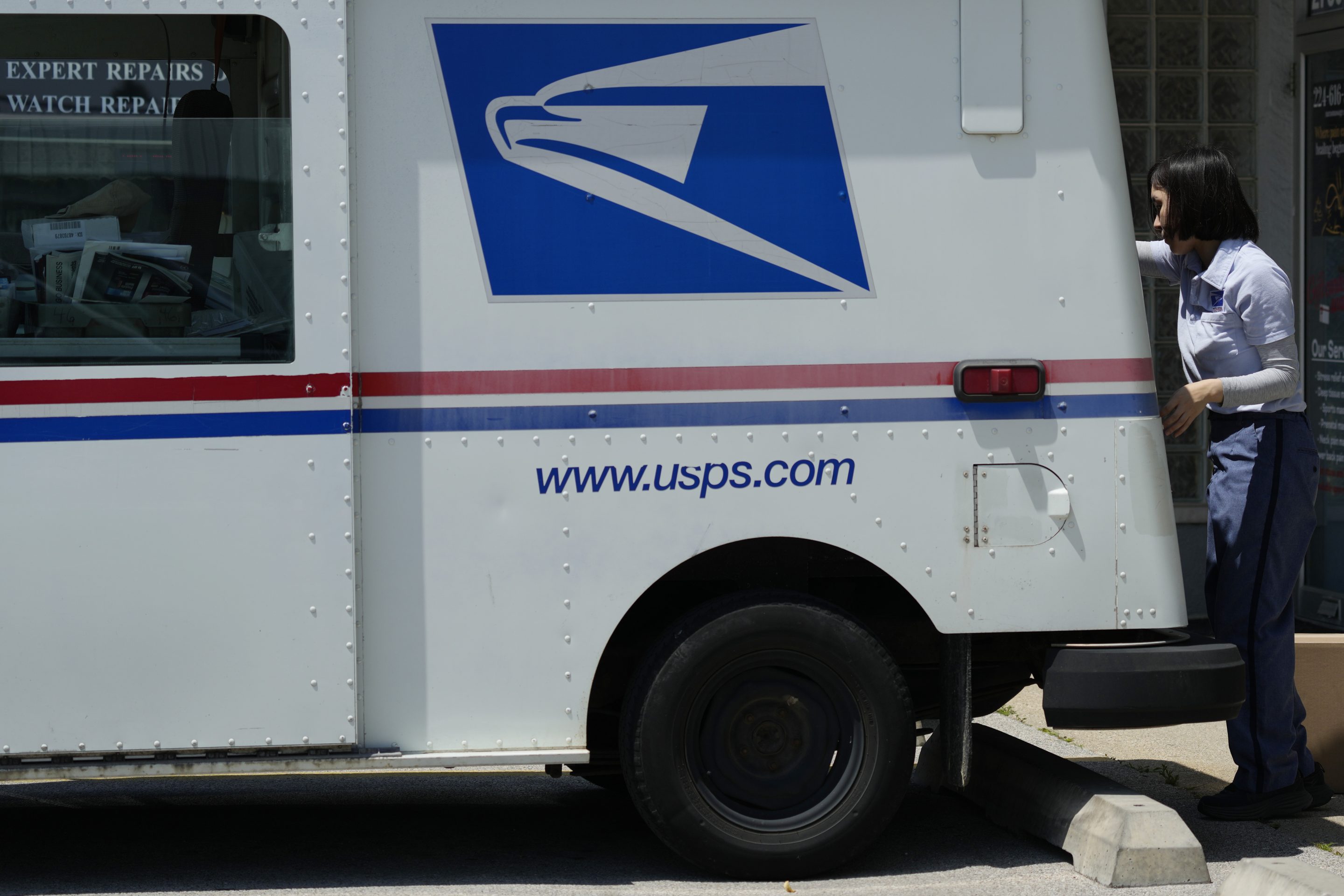 A USPS truck is parked on the sidewalk on a sunny day. The USPS logo is clearly visible and a mail carrier is seen at the back of the truck unloading parcels.