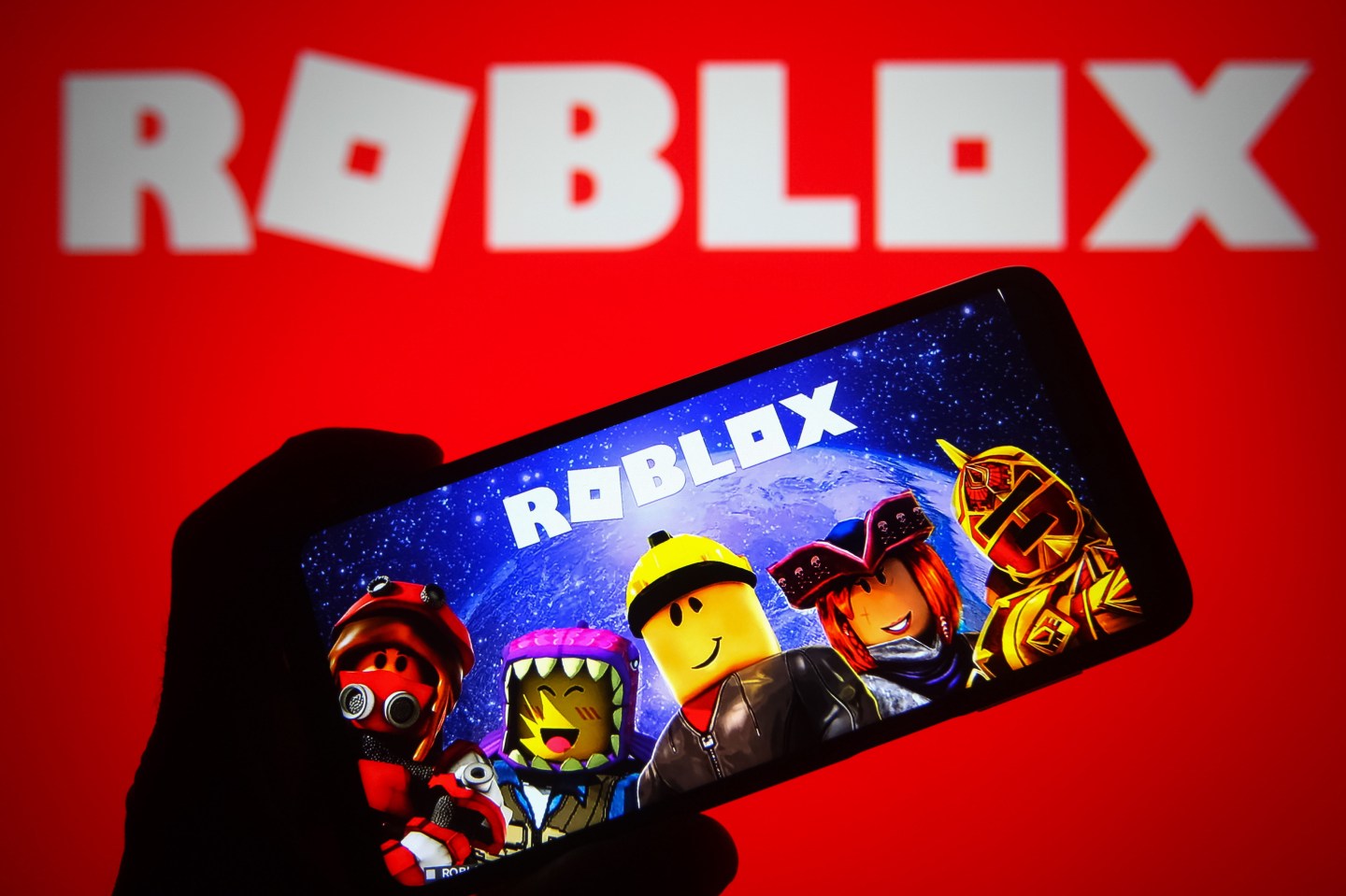 A mobile phone is running the Roblox game app, showing five avatars beneath the Roblox logo.