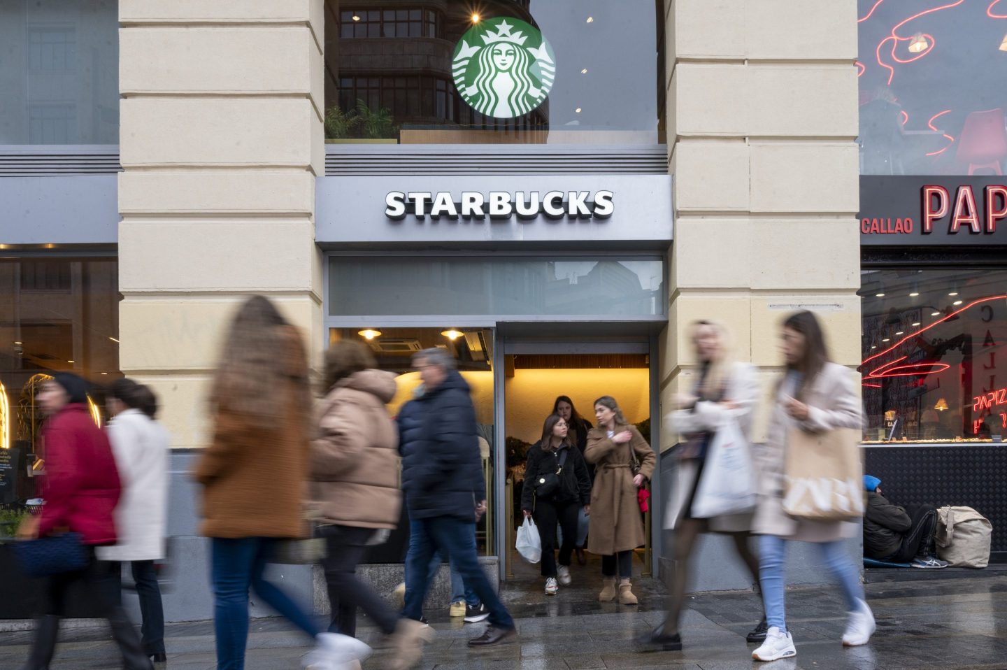 The image shows a busy Starbucks storefront with a crowd of people walking past in both directions. The familiar Starbucks logo with the mermaid is prominently displayed above the entrance. Some people are exiting the store while others are walking by on the street, with a mix of blurred motion capturing the bustling urban environment. The setting appears to be in a city, with a nearby brightly lit shop visible in the background.