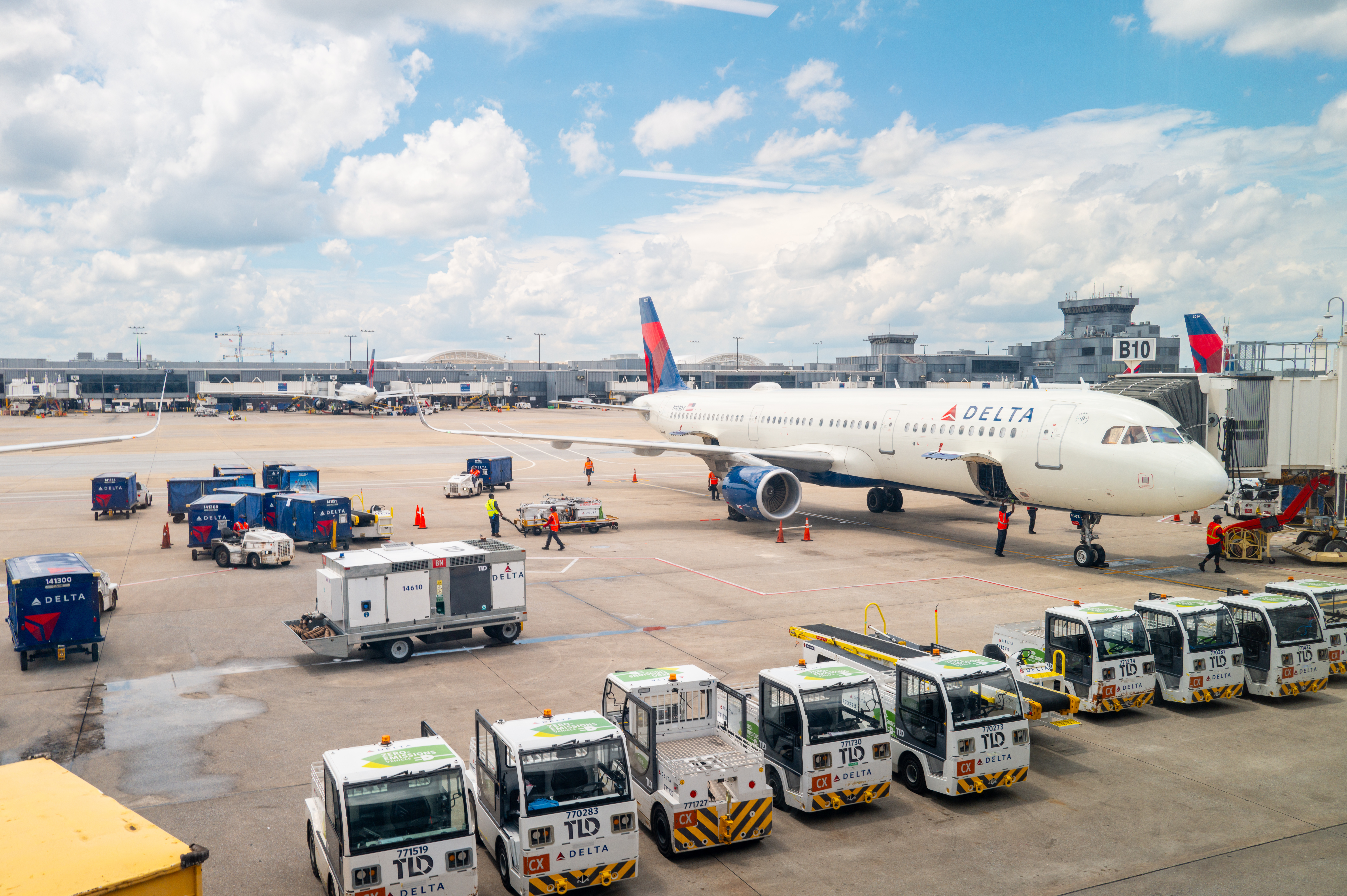 Delta Airlines aircraft parked at a busy airport gate, with ground crew working to load luggage and prepare the plane for departure. Several baggage carts and ground service vehicles are visible on the tarmac under a bright, partly cloudy sky.