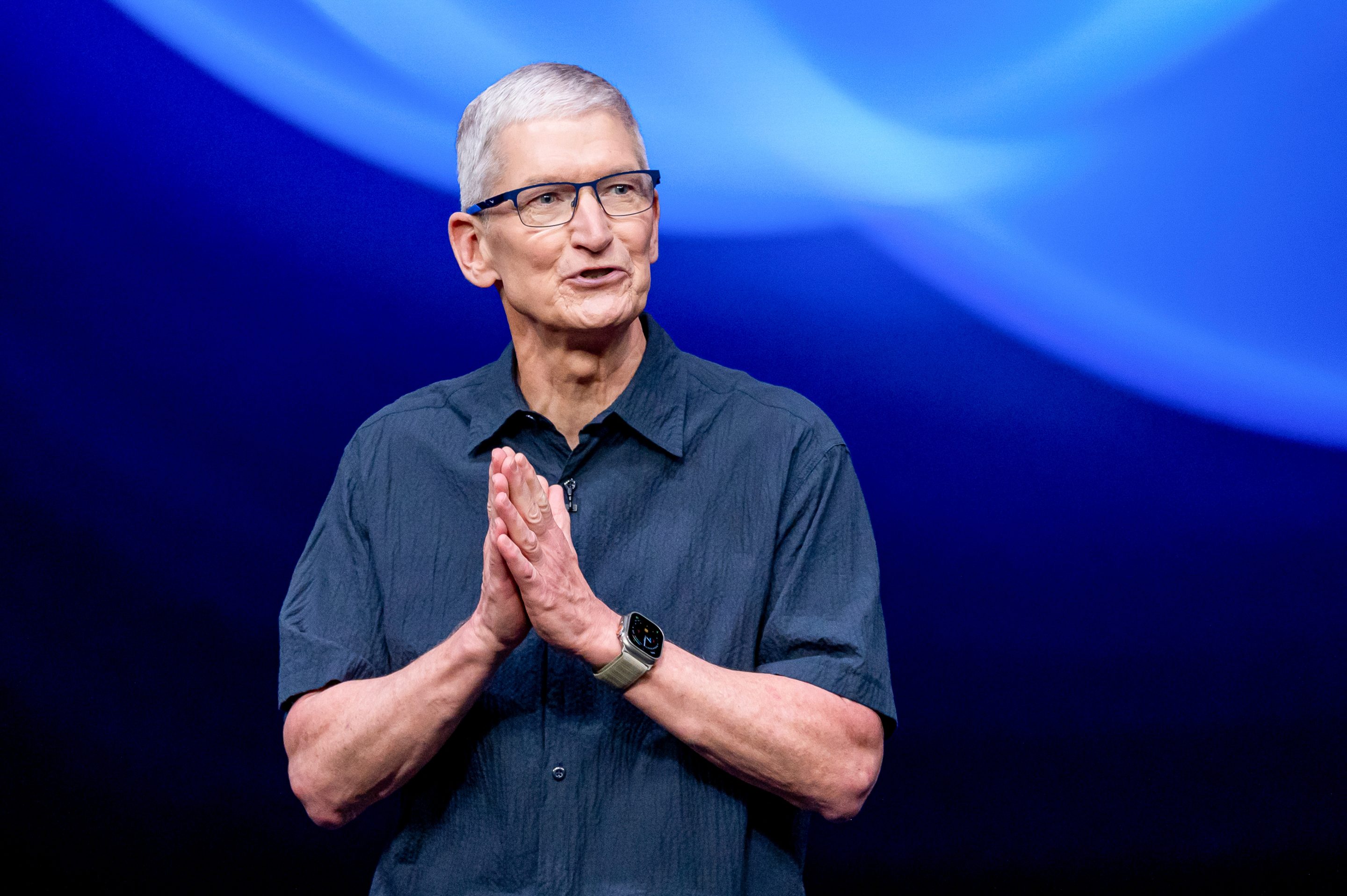 Apple CEO Tim Cook, dressed in a blue button-down shirt, smiles and puts his palms together as he speaks on stage.