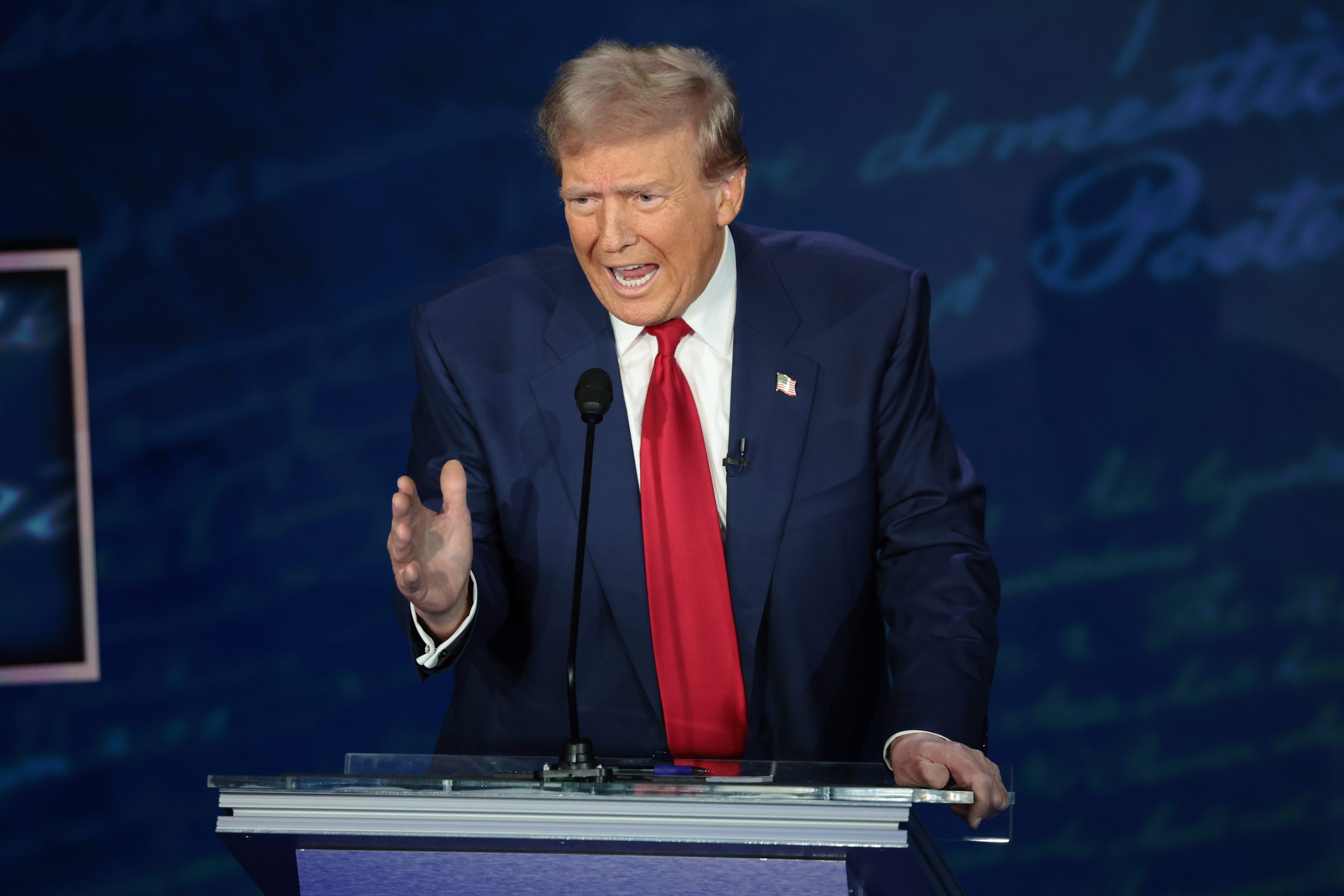 Former President Trump is seen behind the podium at his debate with Vice President Kamala Harris. he is wearing a dark navy suit with a long, bright red tie. He is gesturing with his right hand and his mouth is open as he speaks.