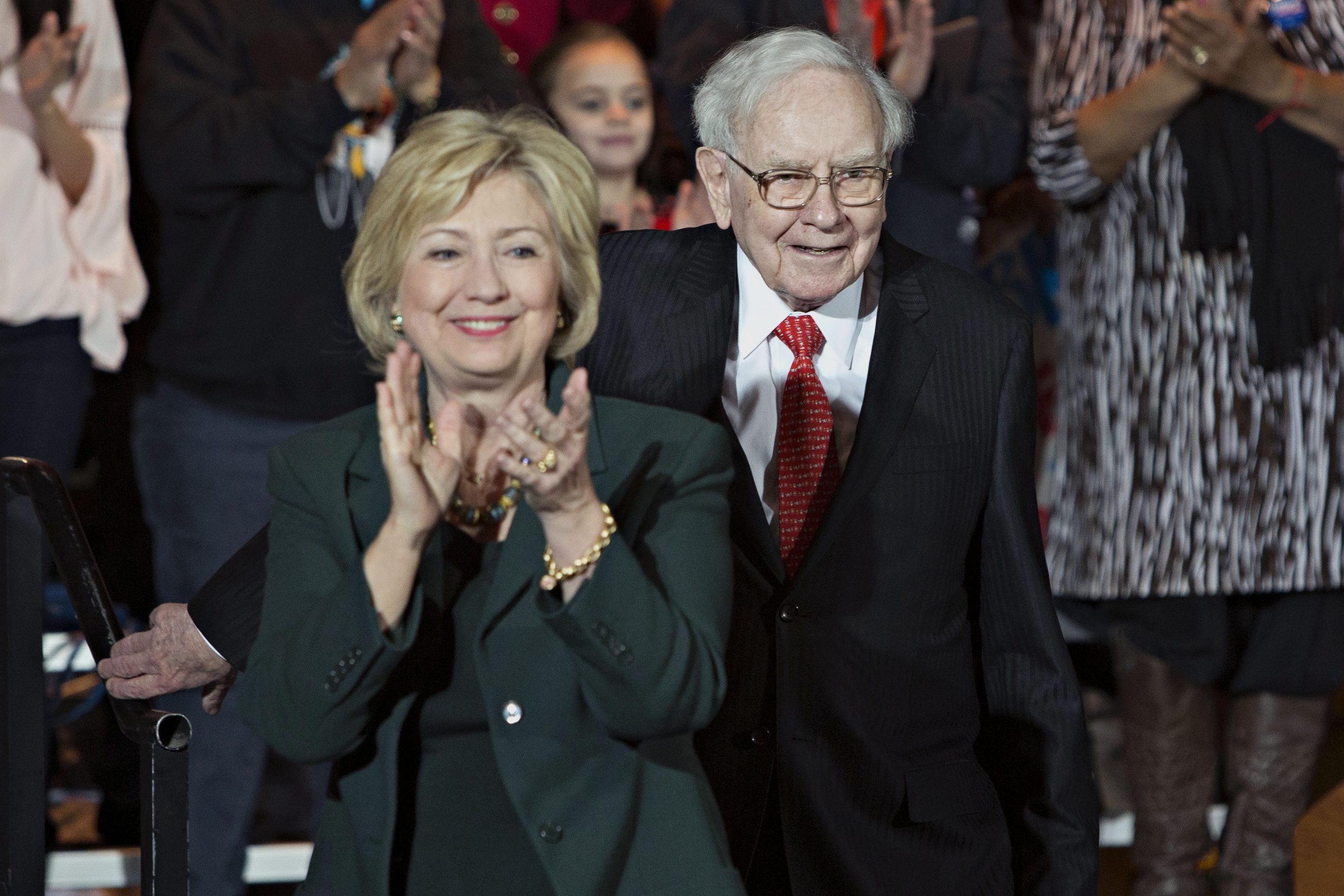 Hillary Clinton, dressed in a green jacket, claps as Warren Buffett, dressed in a black suit, white shirt and red tie, walks behind.