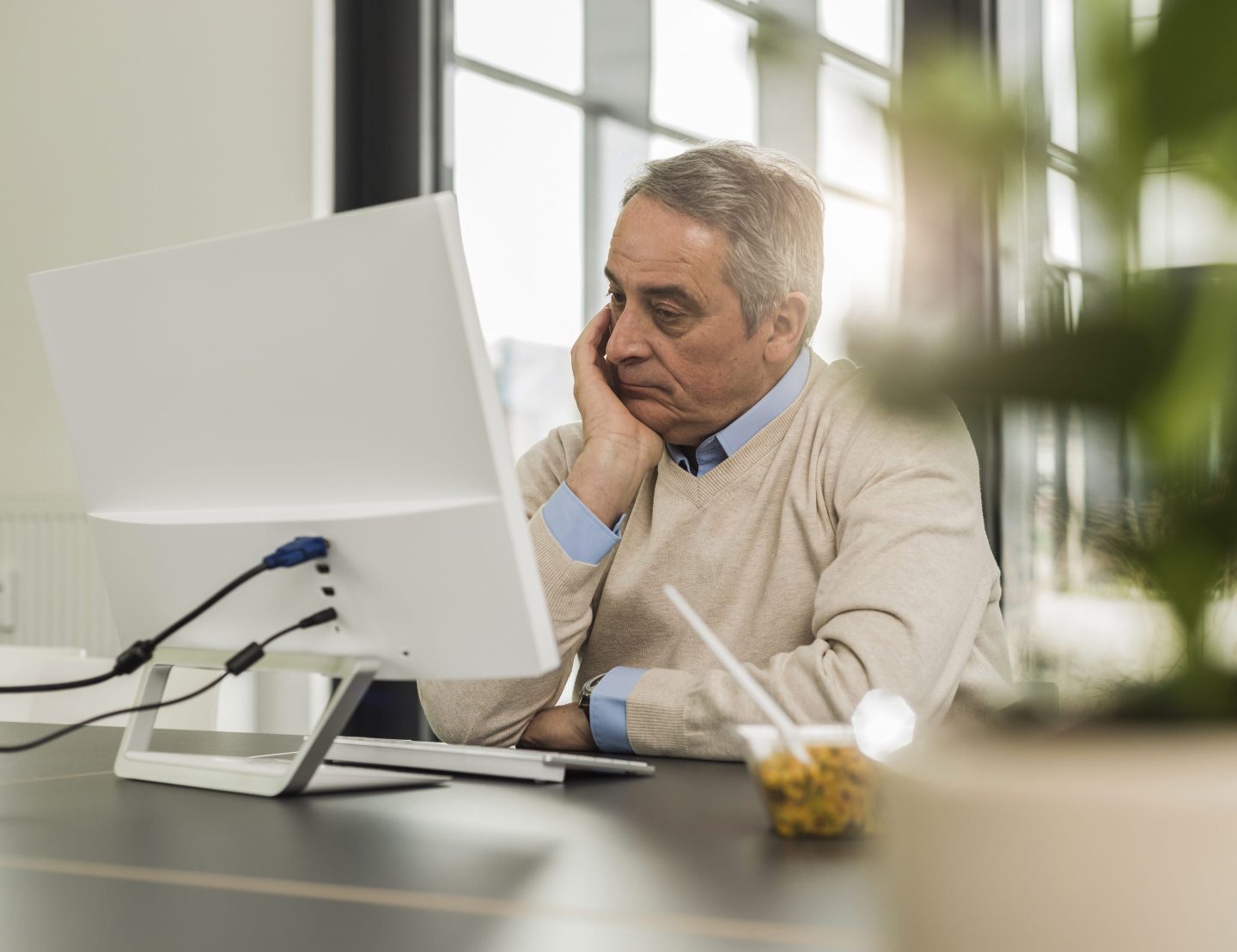 An older man sits in front of a computer with his head in his hand.