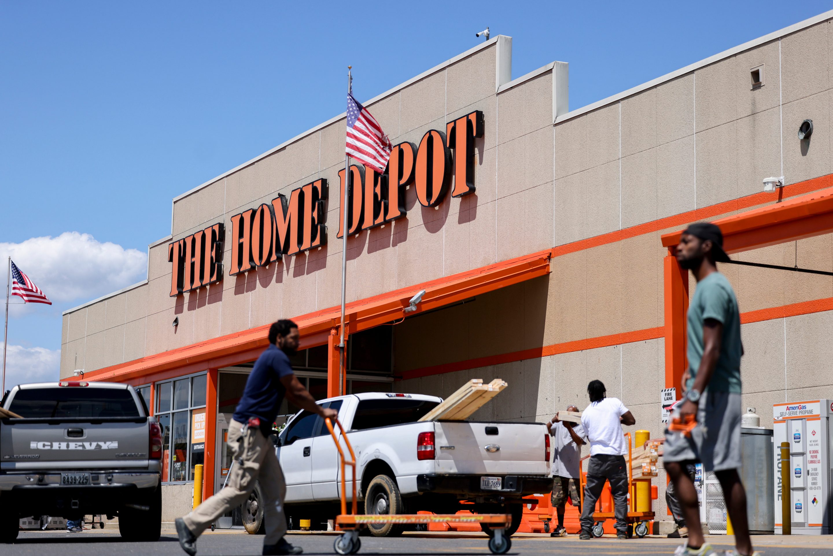 A Home Depot store is seen from the parking lot, with people entering the store and a white truck loading lumber.