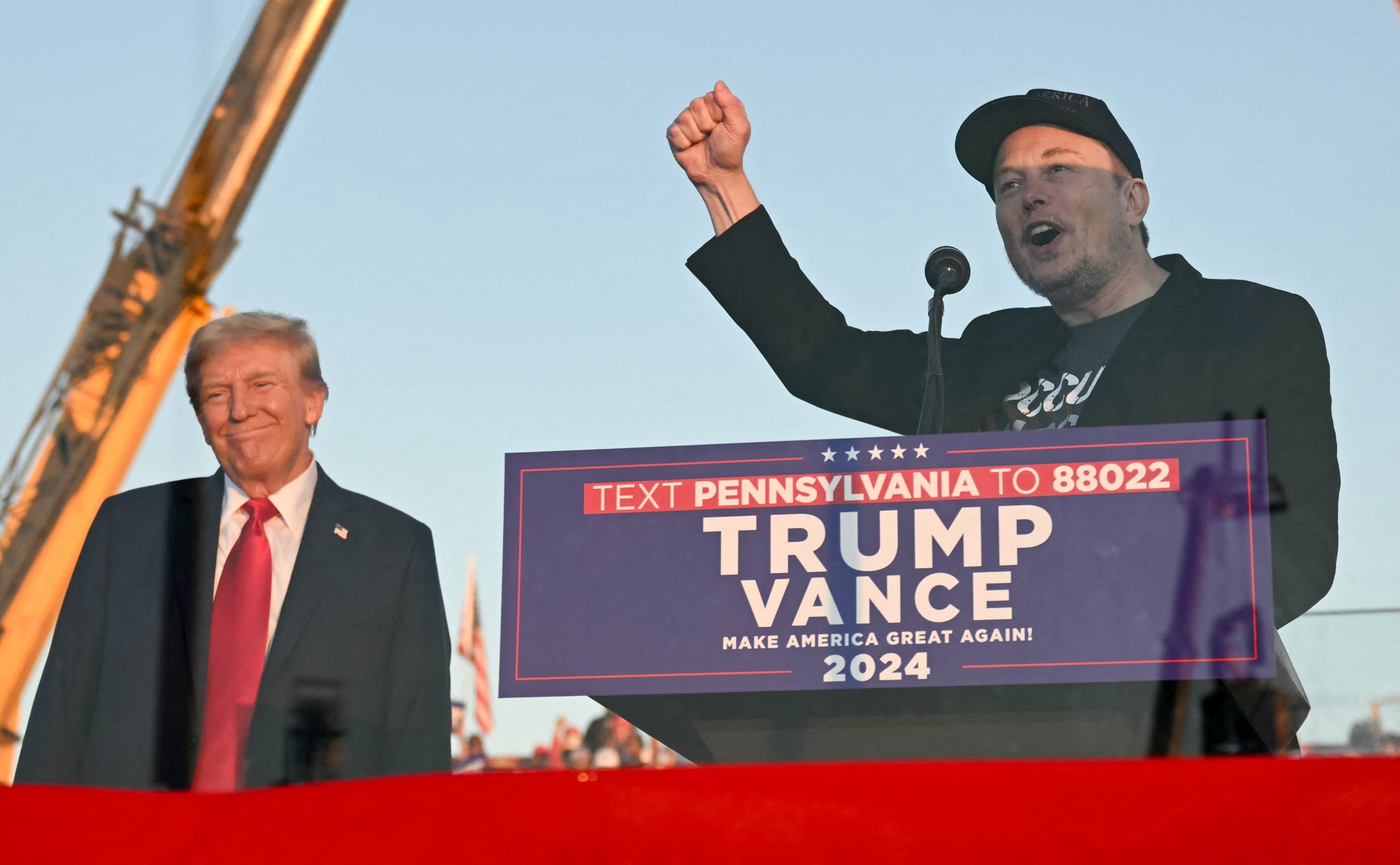Elon Musk raises his first and smiles while speaking at a podium with Donald Trump smiling in the background.