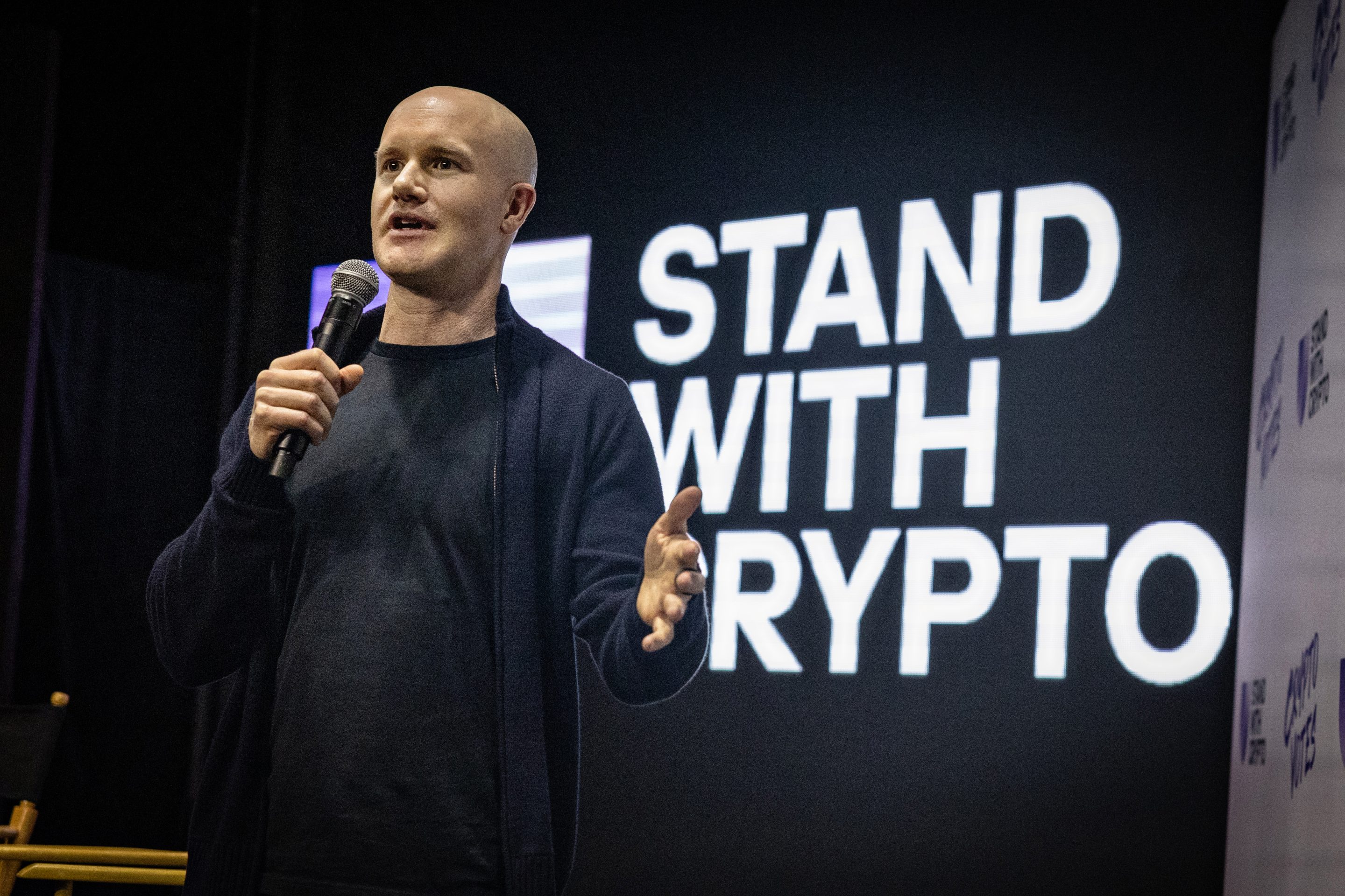 businessman speaks onstage with a "stand with crypto" screen behind him