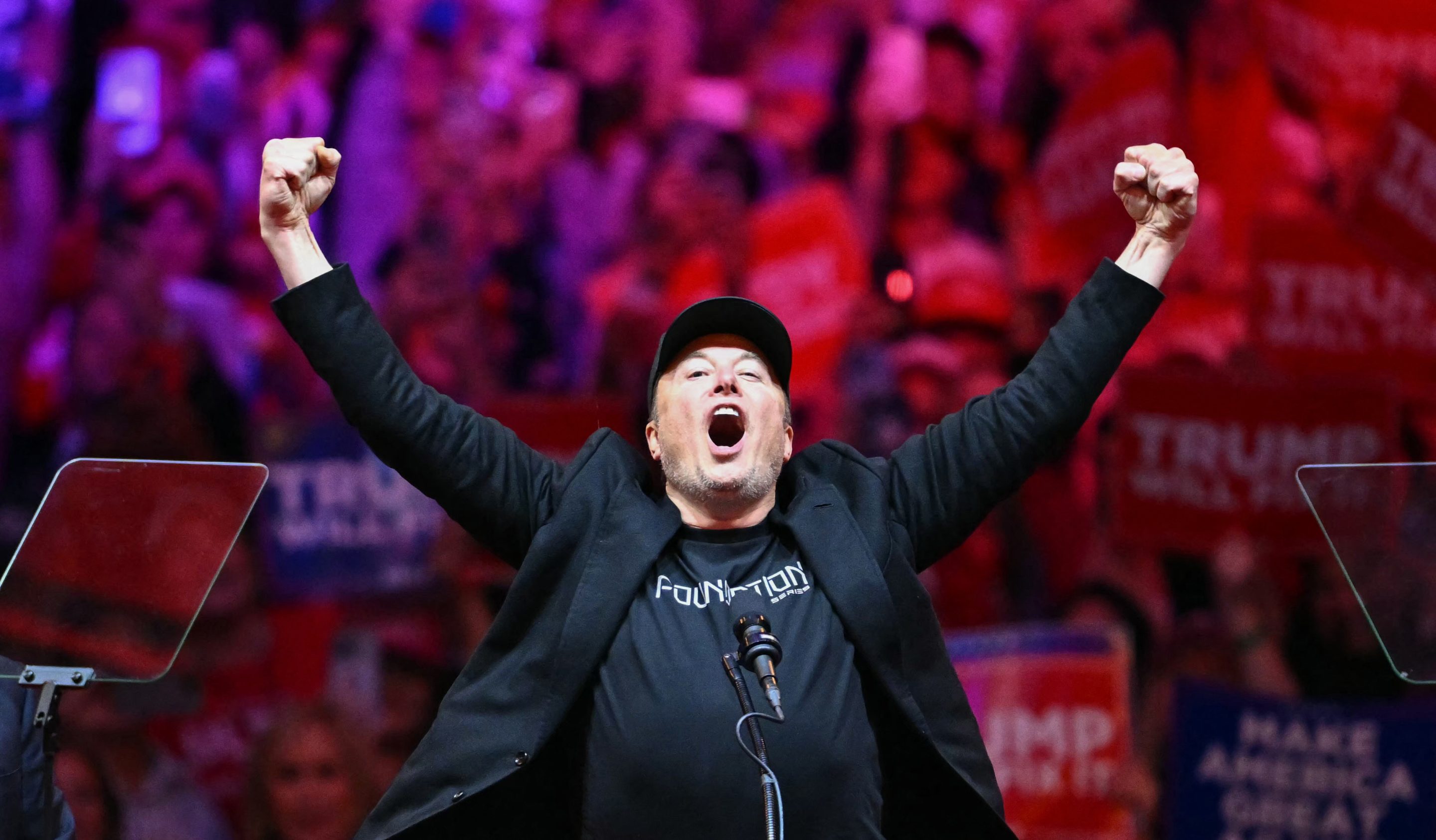 Elon Musk, clothed in black, shouting in front of a microphone with his arms in the air