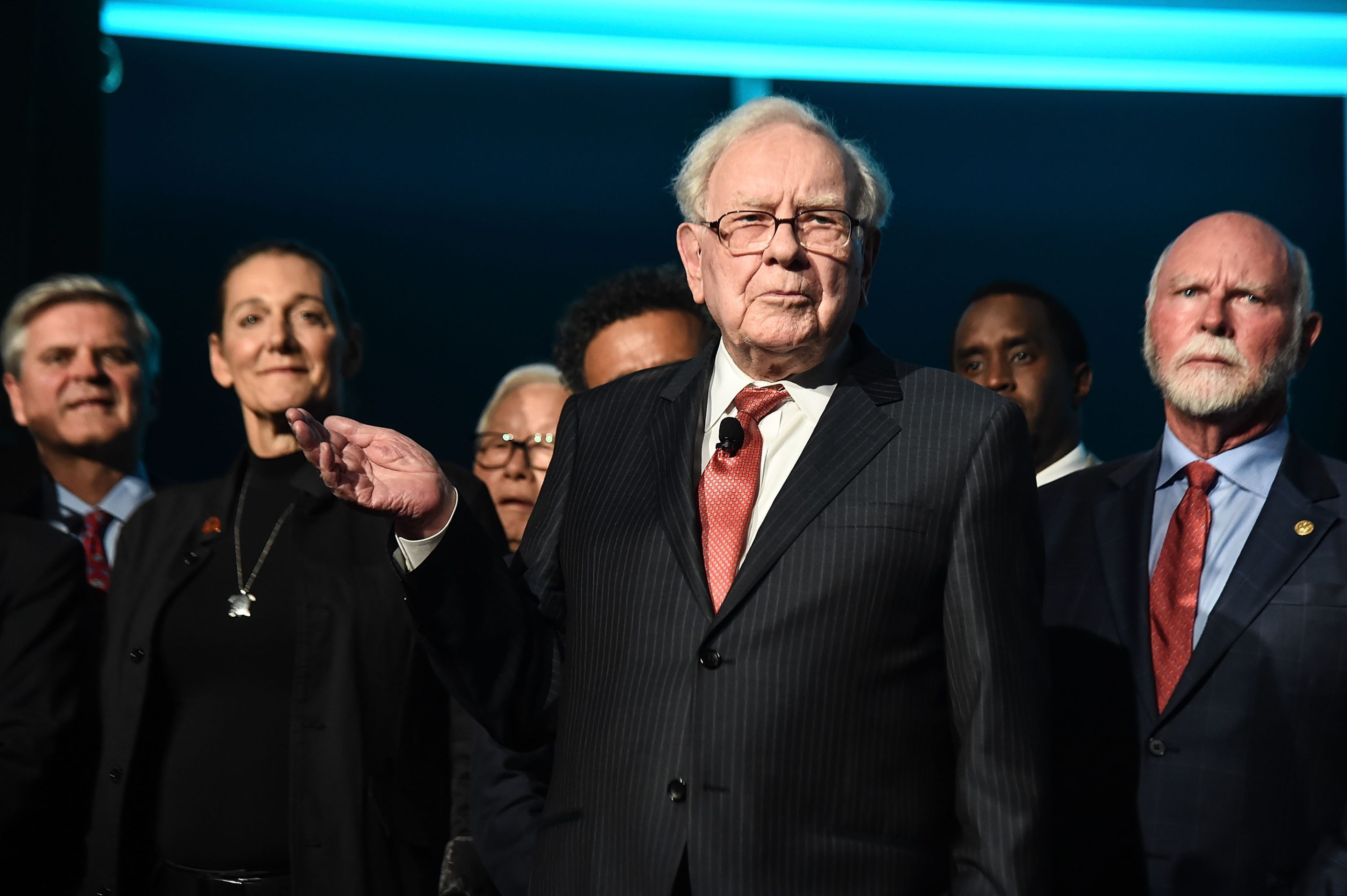 Warren Buffett purses his lips and lifts his right arm as he speaks on stage with other philanthropists and businesspeople on stage in the background.