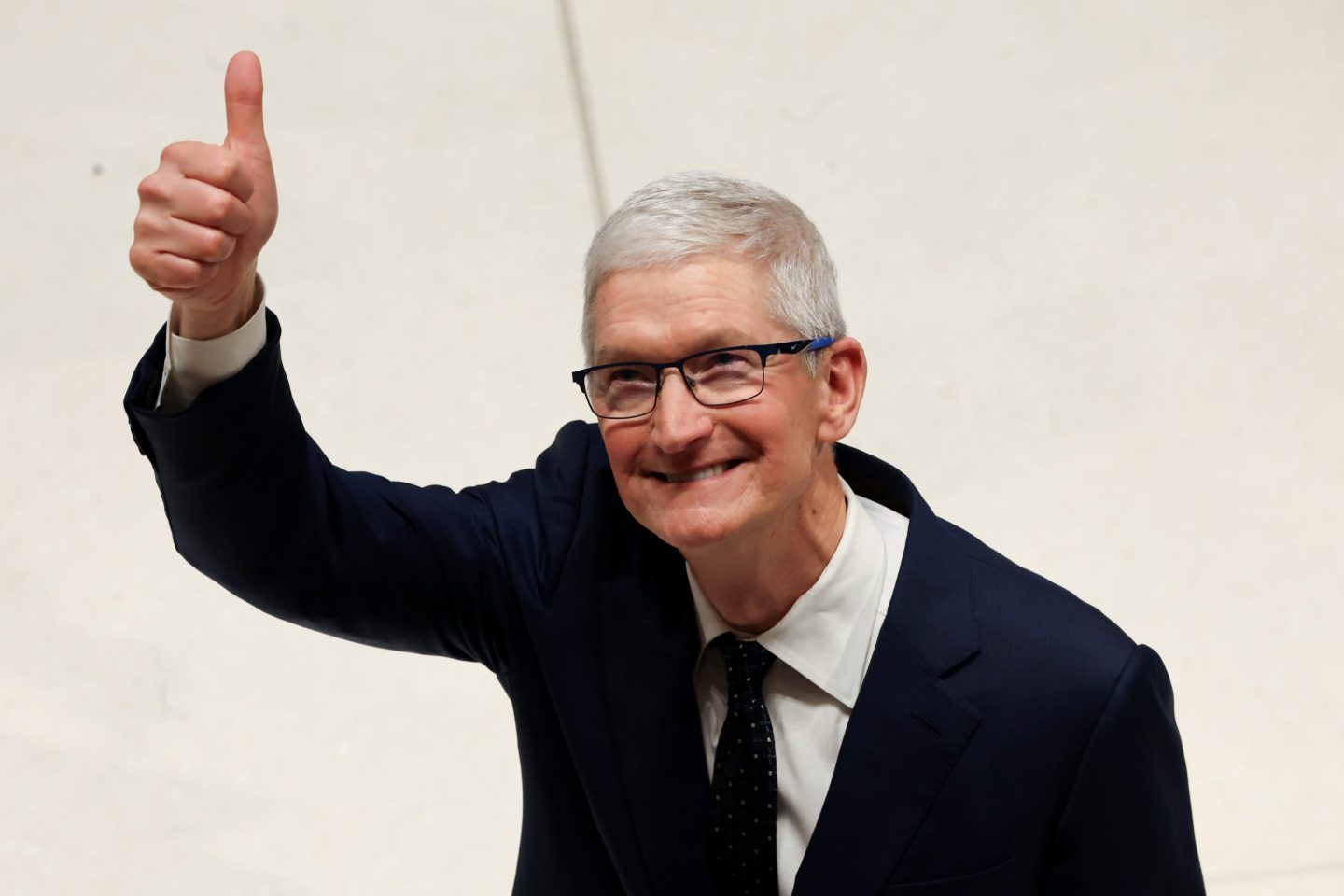 Apple CEO Tim Cook, dressed in a suit and tie, looks up and gives a thumbs up.