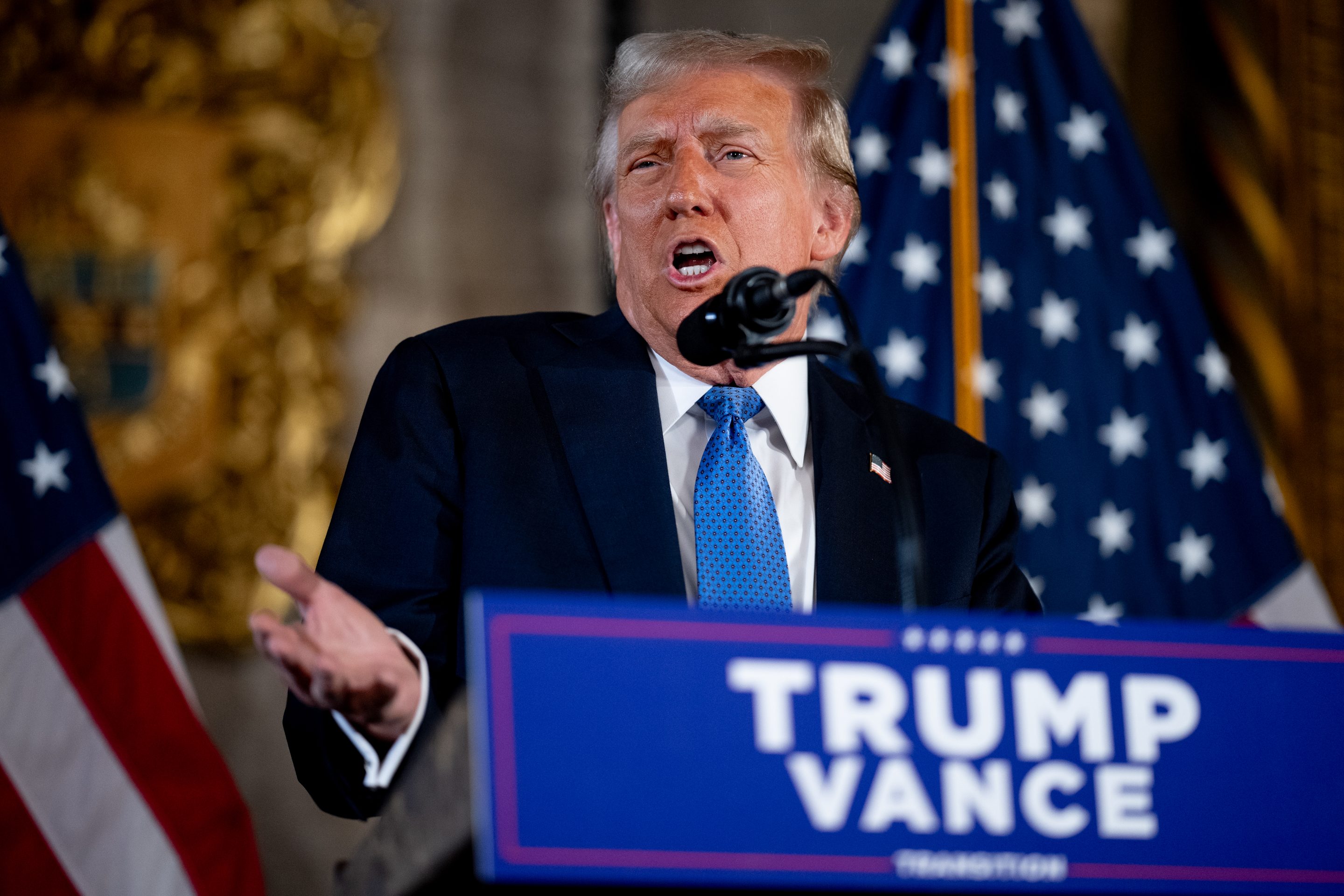 Donald Trump shrugs and gestures as he speaks into a microphone behind a "Trump Vance" podium.