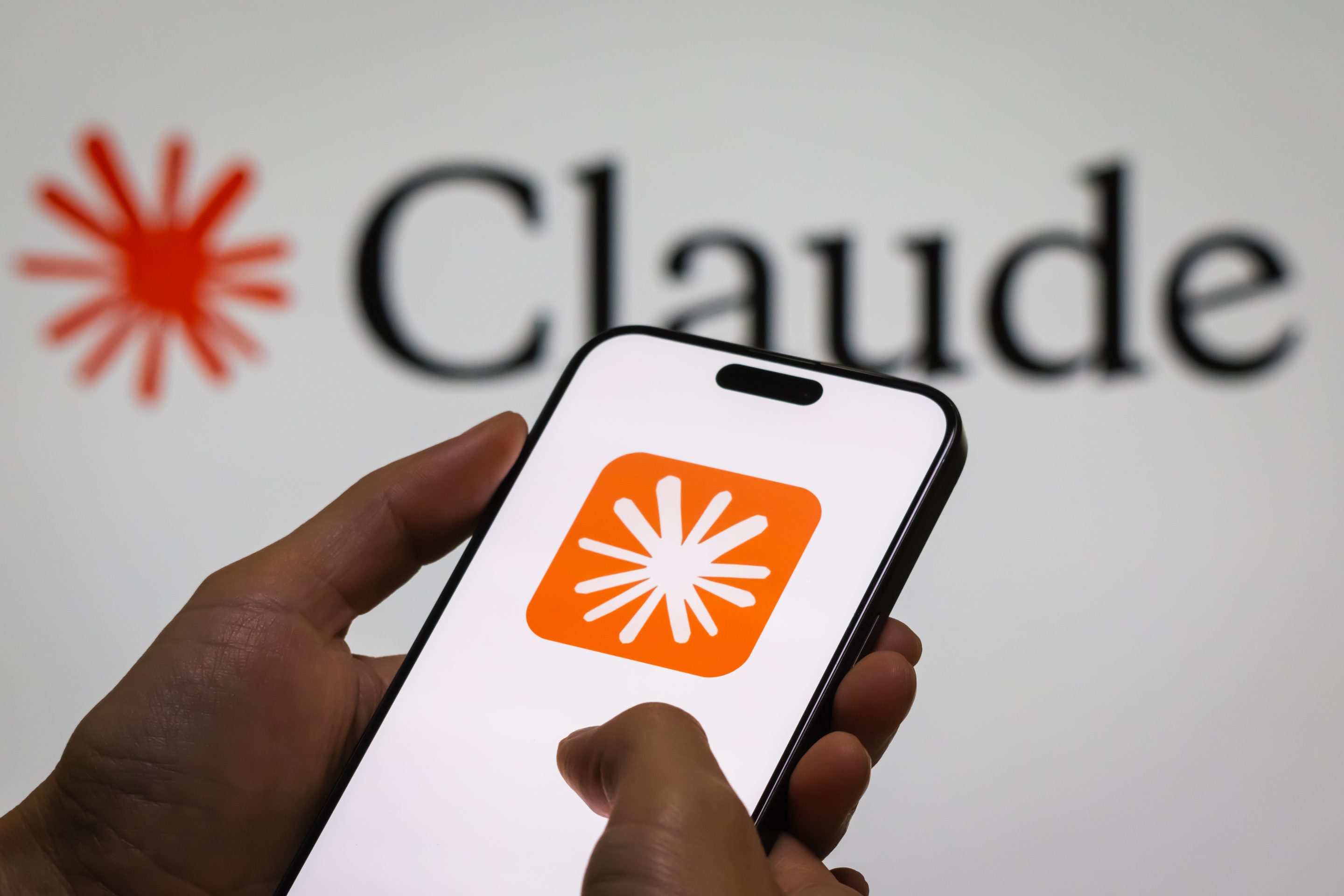 An image of a hand holding a mobile phone displaying the logo of Anthropic's Claude AI chatbot against a backdrop similarly showing the Claude logo and the word "Claude."