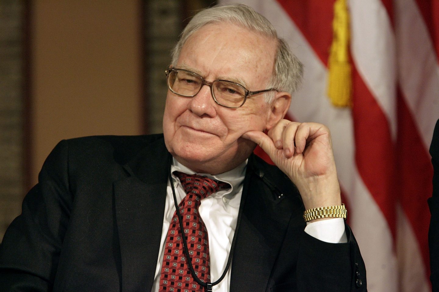 Warren Buffett sits and leans his head on his left hand as he listens with an American flag in the background.