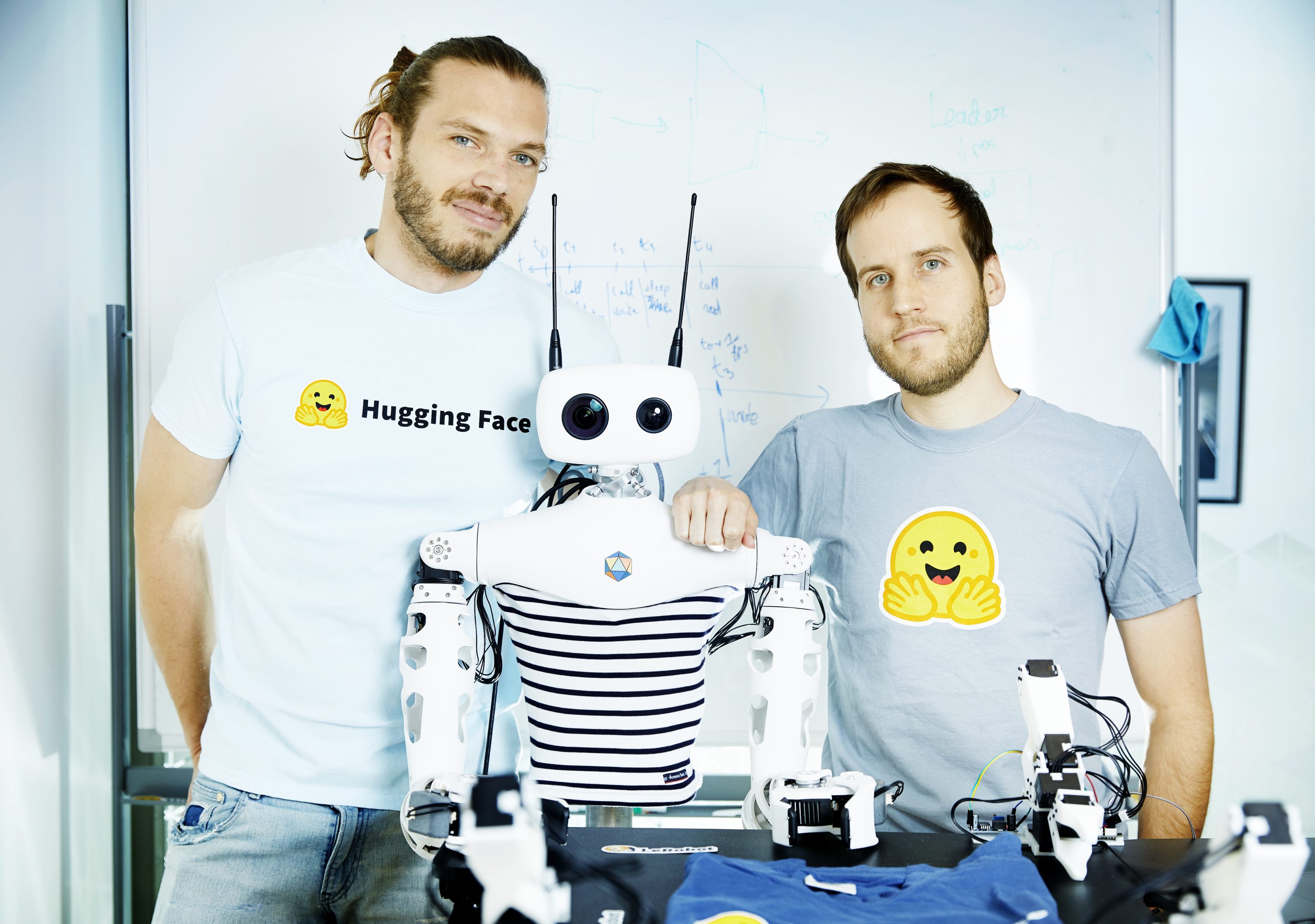 Simon Alibert and Rémi Cadene, robotics researchers at Hugging Face, pose wearing T-shirts with the Hugging Face logo as they stand alongside Reachy 2, a humanoid robot built by Pollen Robotics.
