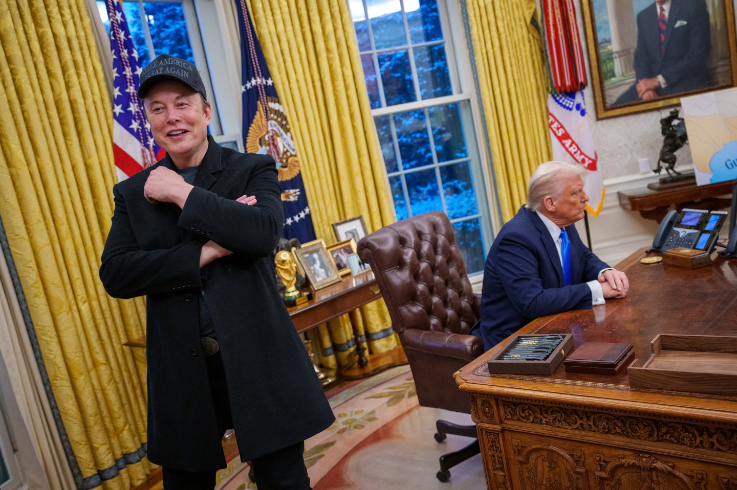 Elon Musk standing in the oval office with Donald Trump sat behind.