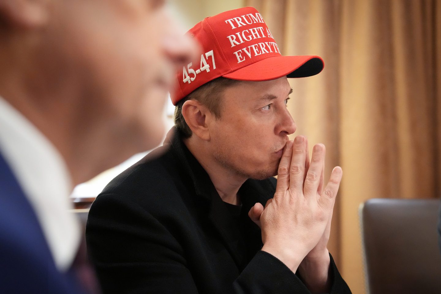 Elon Musk, wearing a MAGA hat, puts his fingers to his lips and he looks forward.