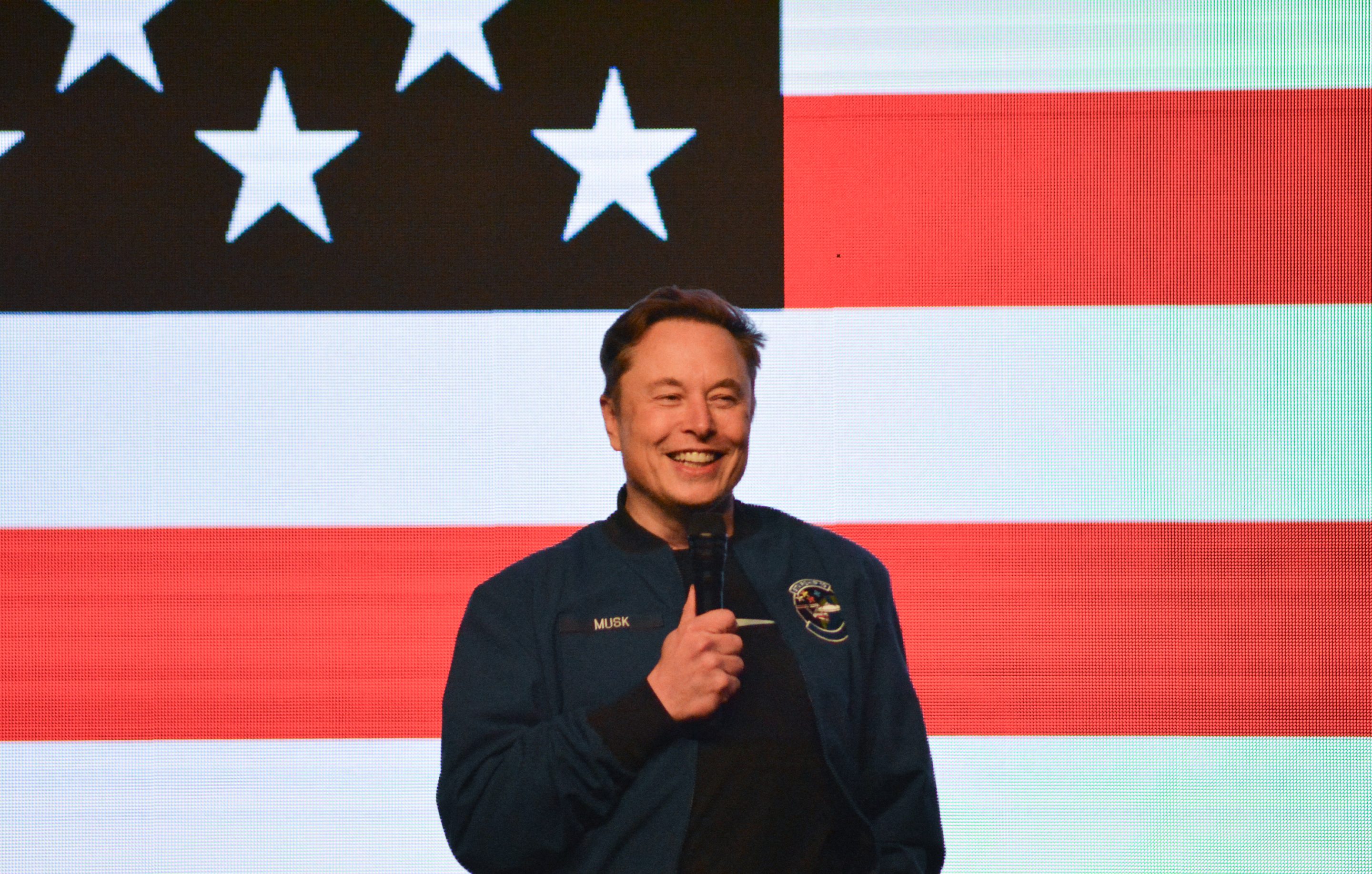 Elon Musk holds a microphone on stage in front of an American flag