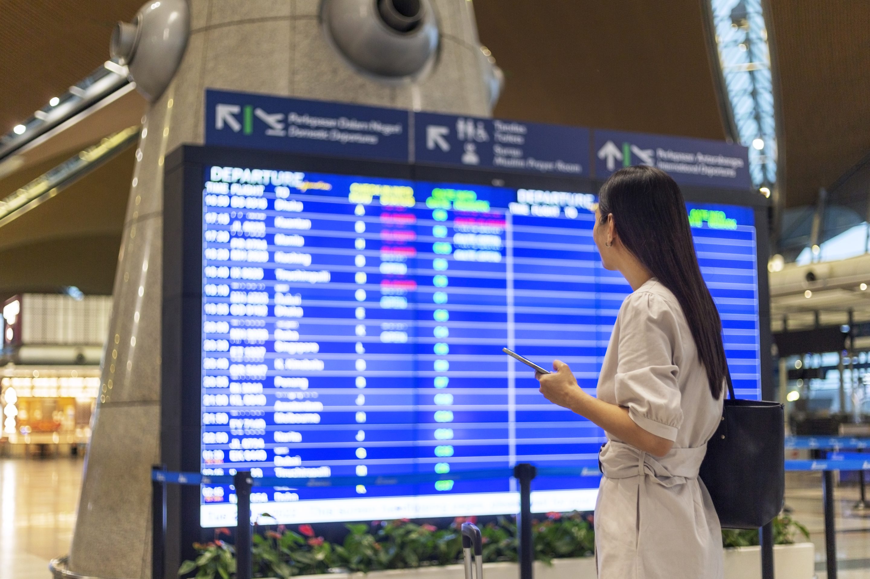 woman with luggage and smart phone checking flight schedule near departure board in airport
