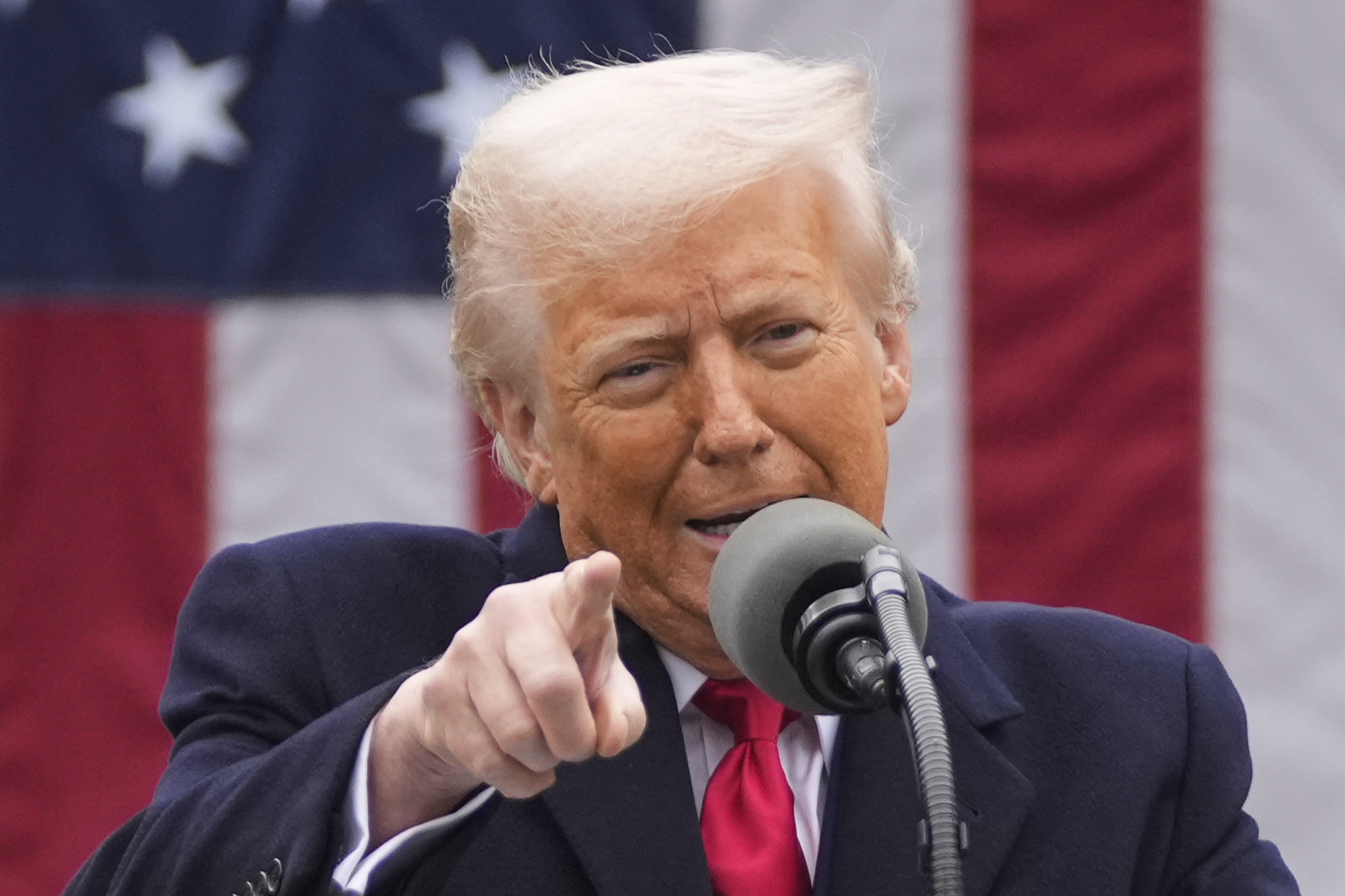 Trump speaks into a microphone and points into the crowd in front of an American flag