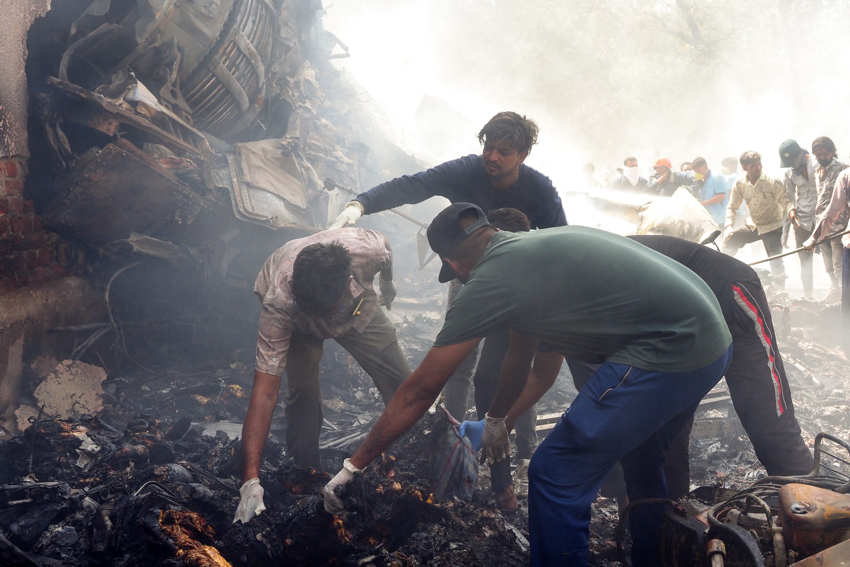 Three people pick up wreckage next to a crashed jet, surrounded by smoke.