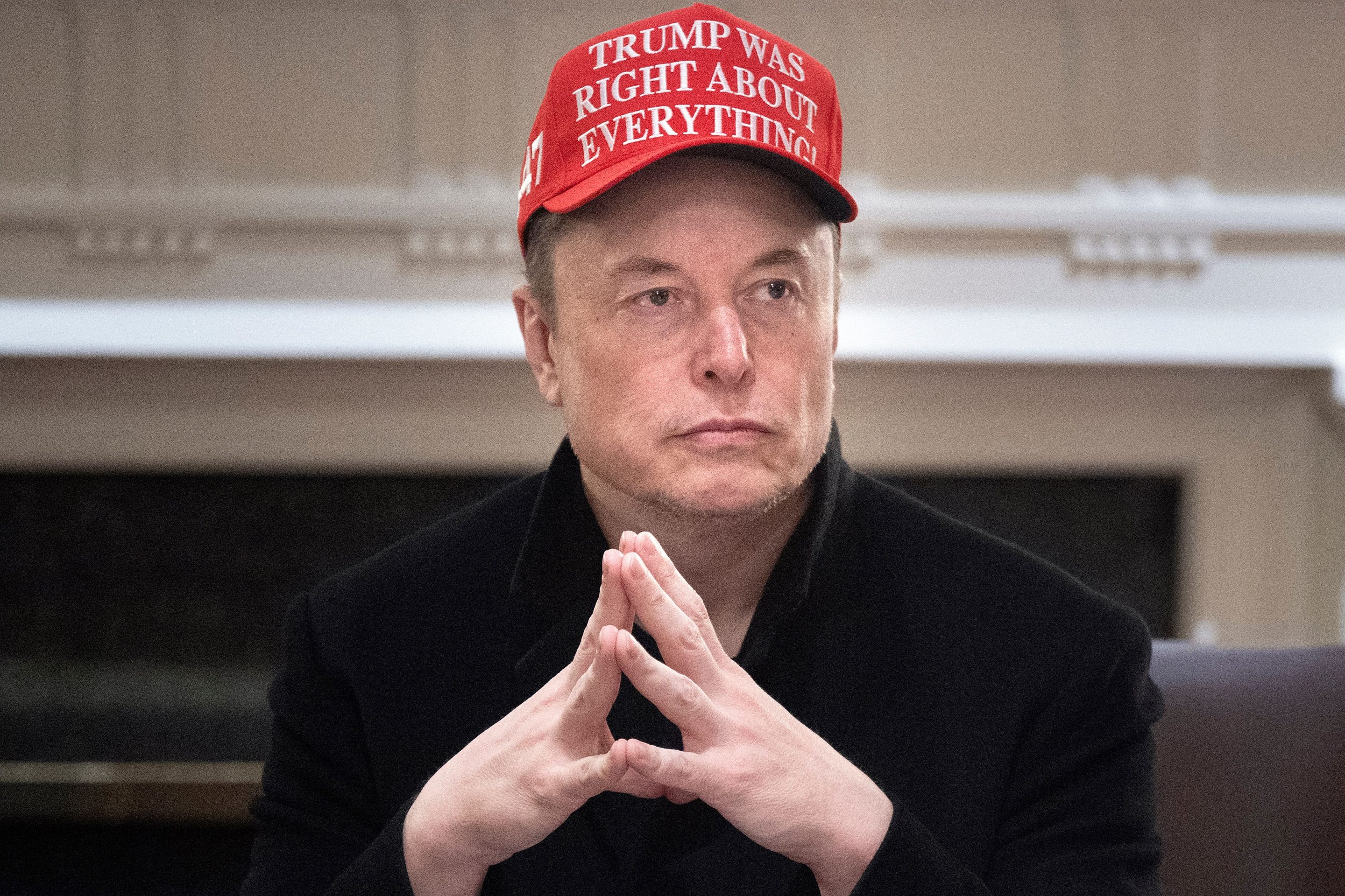 Elon Musk sits with his hands folded wearing a MAGA style hat