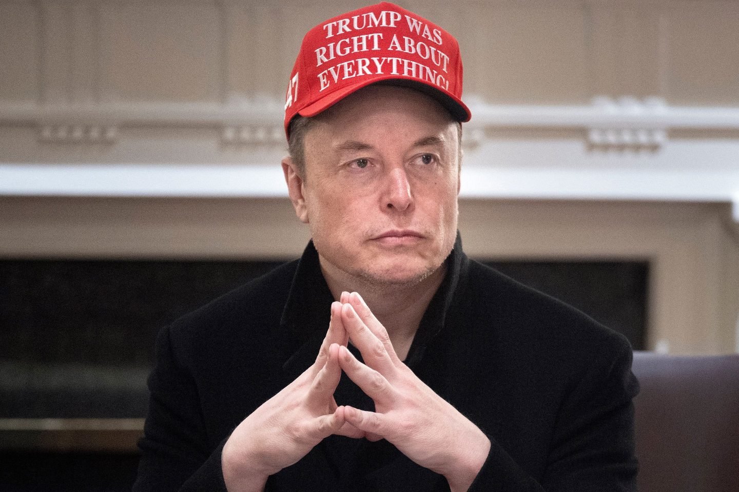 Elon Musk sits with his hands folded wearing a MAGA style hat