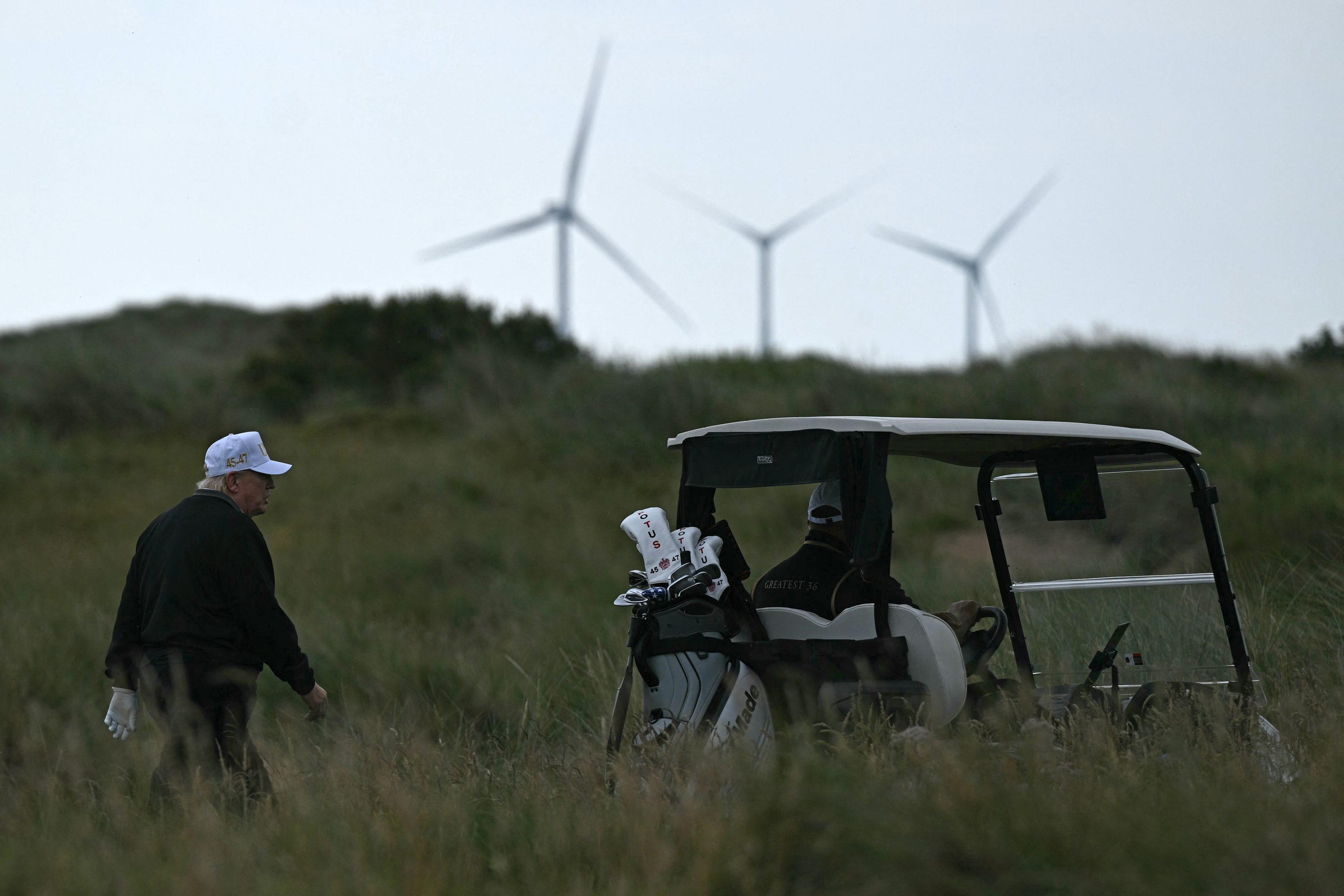 US President Donald Trump (L), backdropped by Turbines at the European Offshore Wind Deployment Centre, also known as the Aberdeen Bay Wind Farm, walks on the first fairway after playing off the first tee to officially open the Trump International Golf Links course in Balmedie, Aberdeenshire, north east Scotland on July 29, 2025.