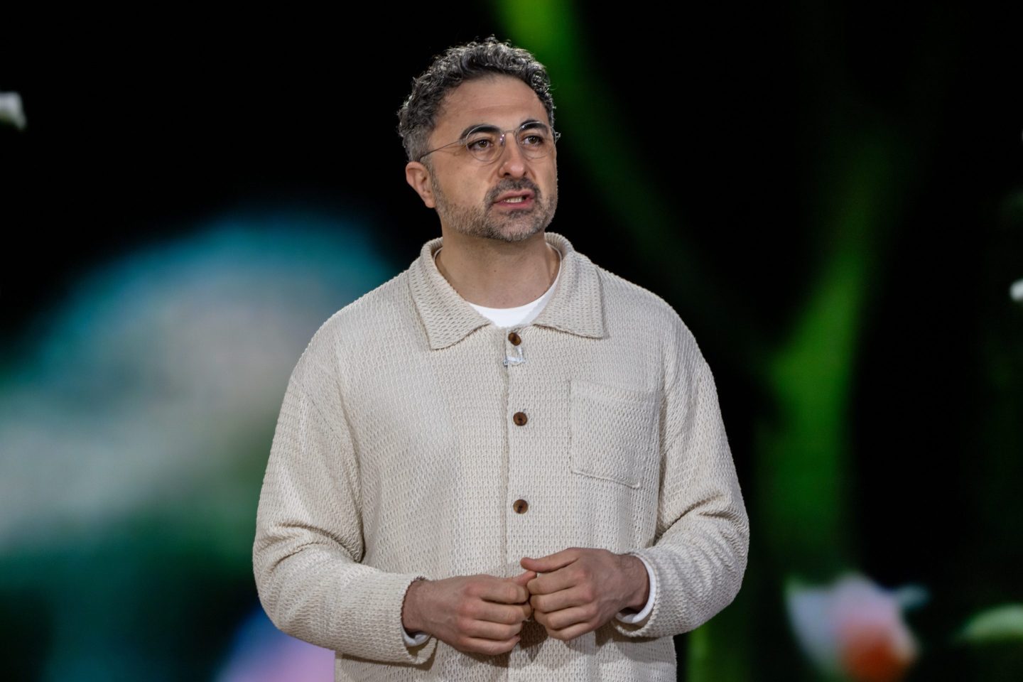 Mustafa Suleyman , wearing a light chore jacket, speaks with both hands in light fists in front of him.