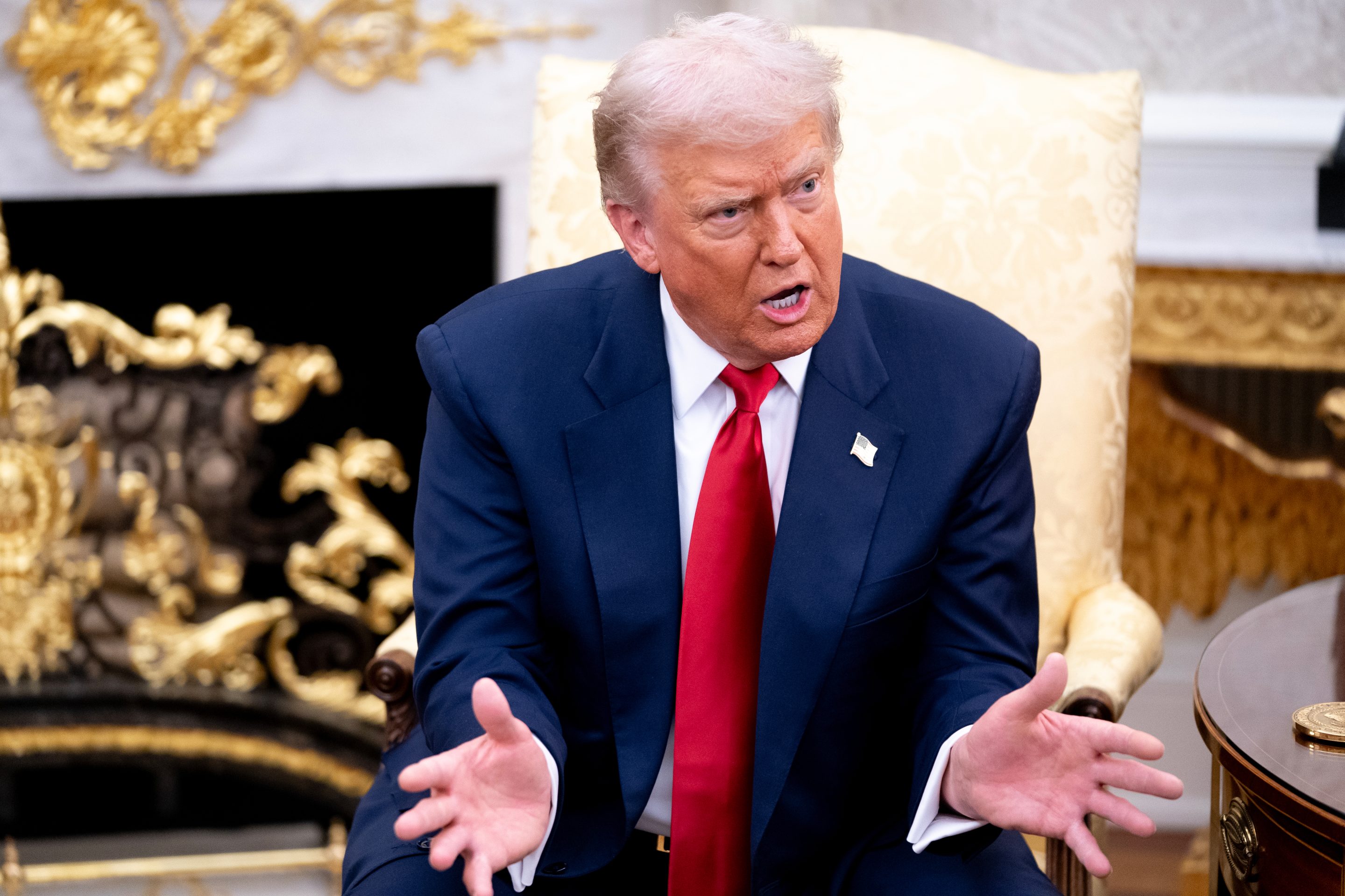 Trump sits in a chair in the Oval Office, speaking while gesturing with both hands.