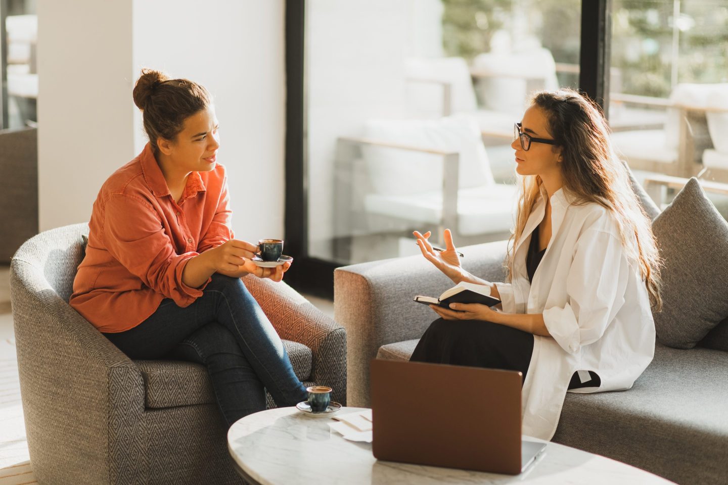 Two women sit in an office talking over coffee.