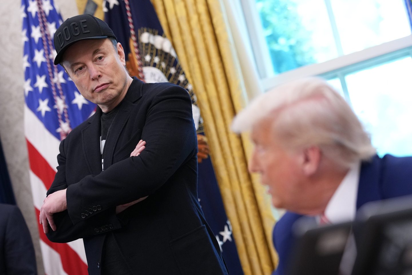 Elon Musk, standing with his arms crossed, looks down at Donald Trump sitting at his desk in the Oval Office