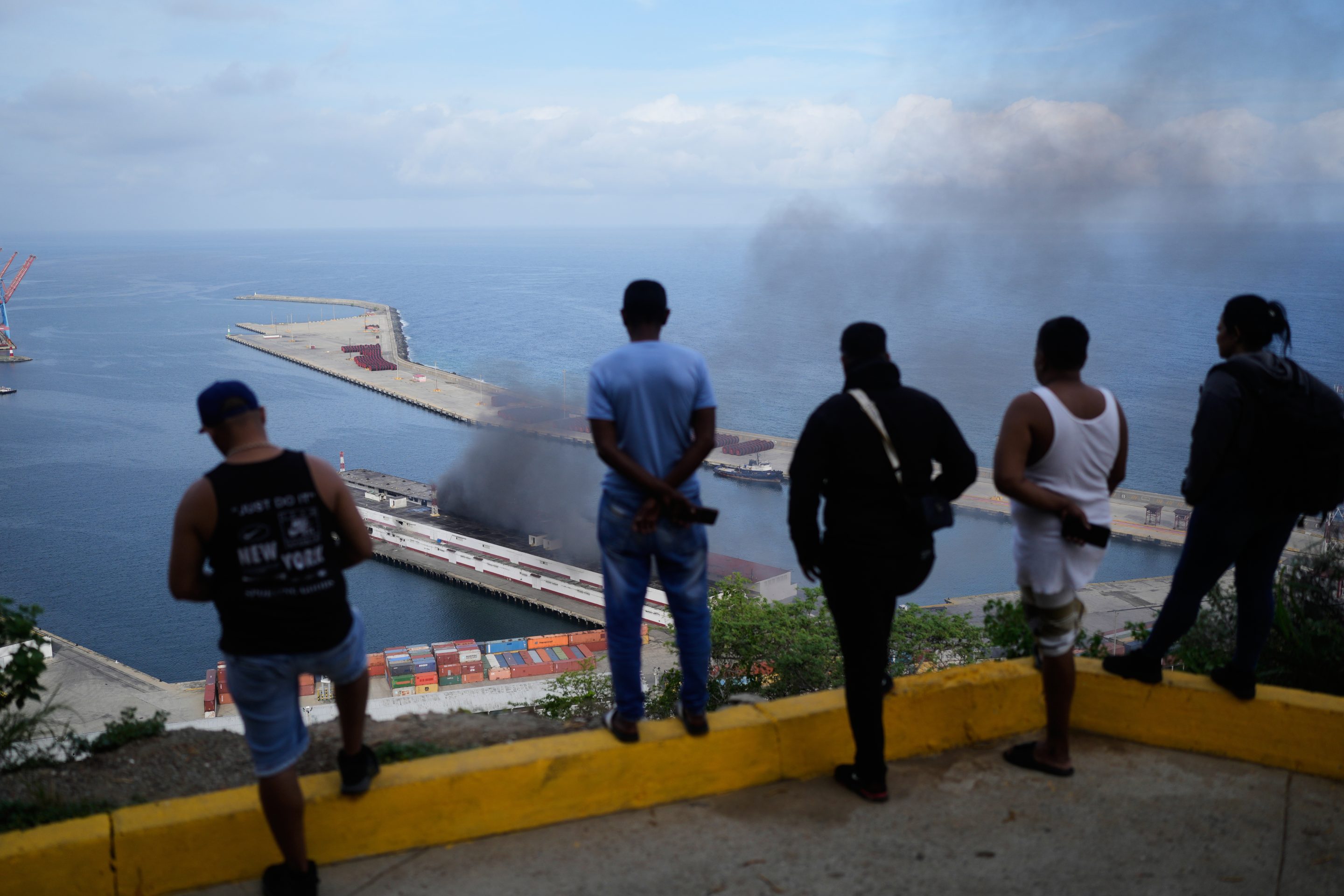 Men watch smoke rising from a dock after explosions were heard at La Guaira port, Venezuela, Saturday, Jan. 3, 2026.