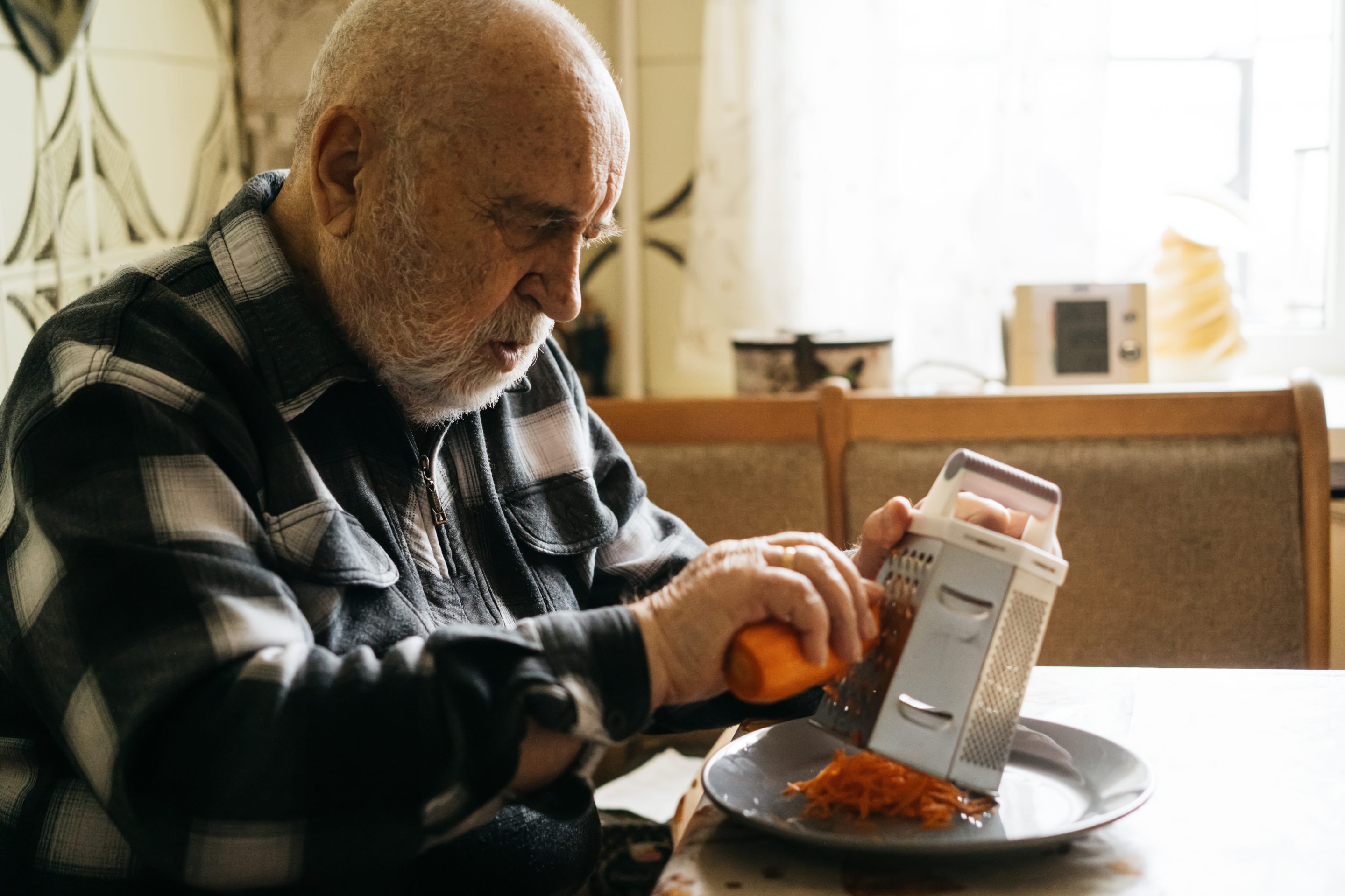 An elderly man prepares ingredients, grating carrots on a plate in a home setting, emphasizing independence and routine.