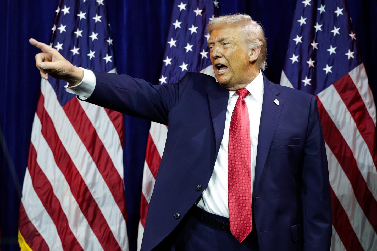 President Trump, standing in front of American flags, points to his right with his mouth open.