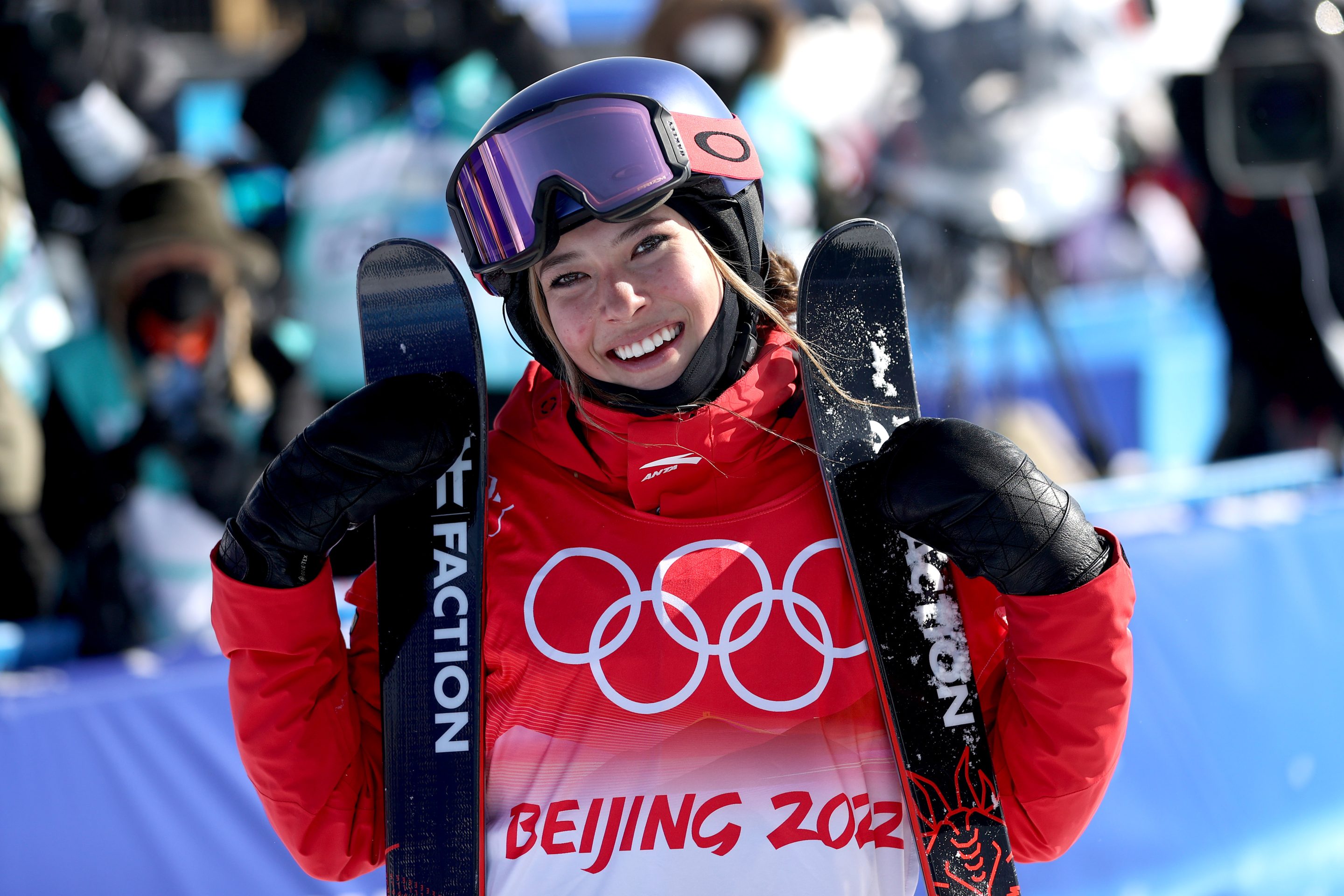 Eileen GU, wearing a red and while Beijing Olympics coat, smiles with her skis.