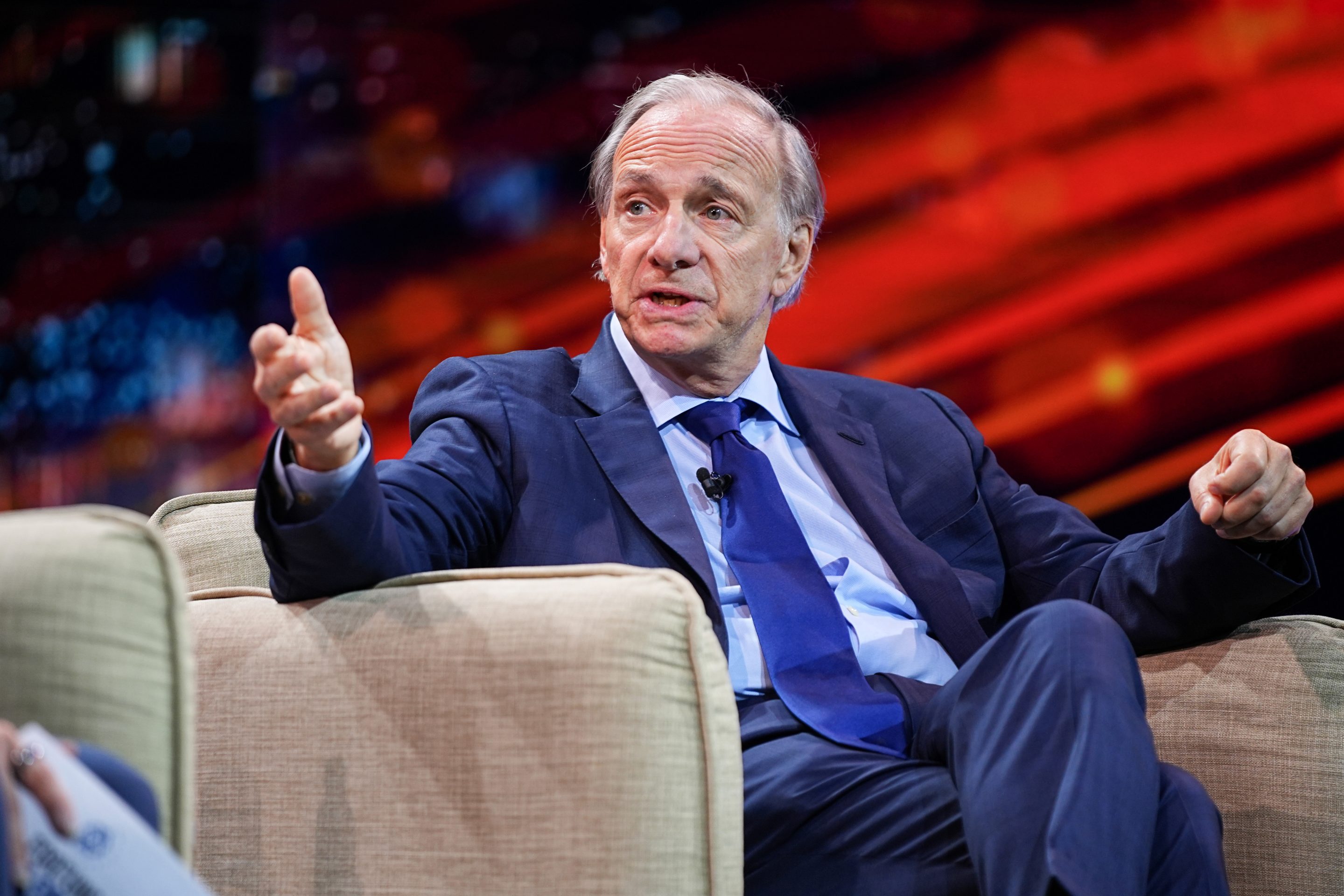 Ray Dalio, wearing a suit and sitting in a beige chair, speaks and gestures with his hand.