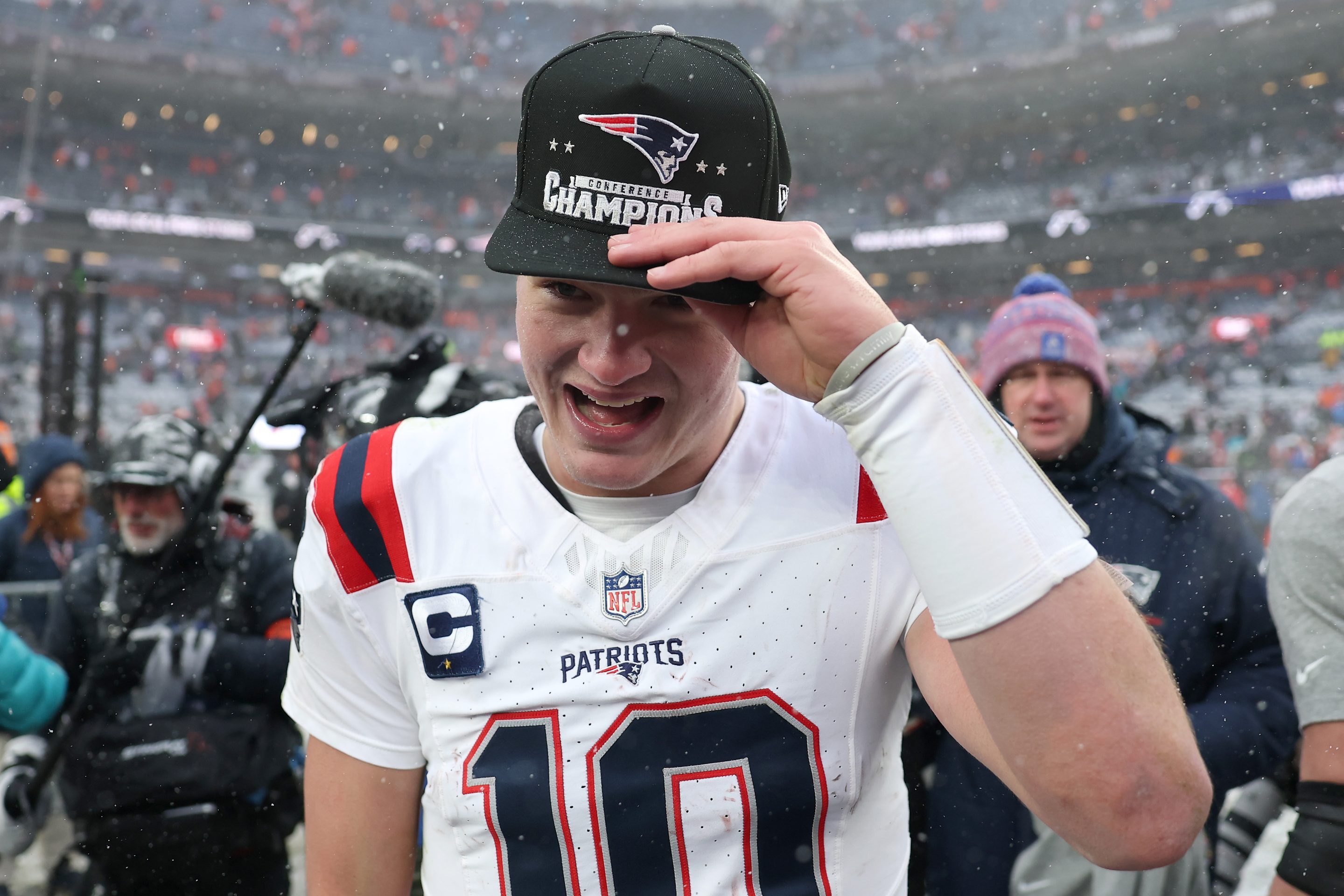 Drake Maye holds onto an AFC Conference Champion hat as he smiles on the field.
