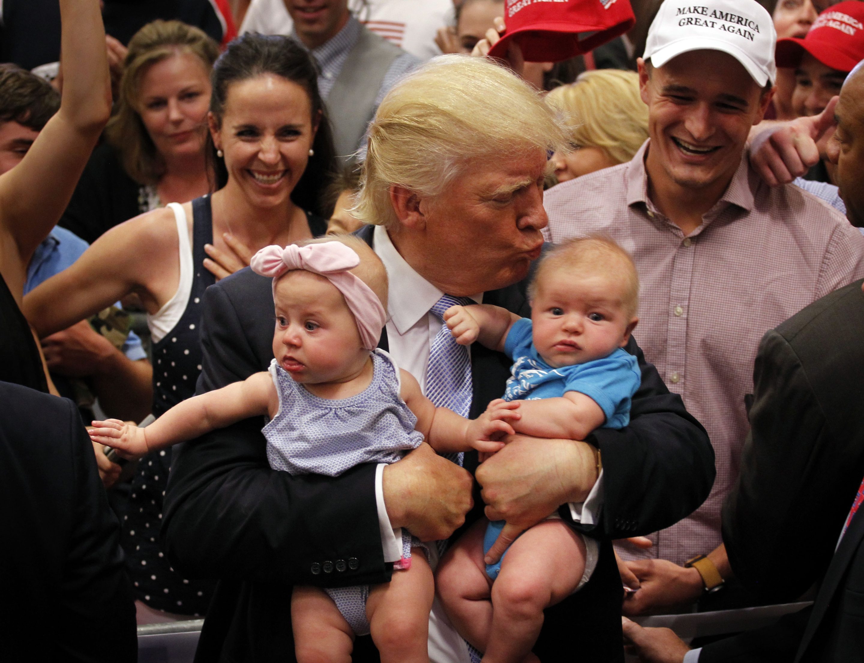 Donald Trump, holding two babies in his arms, leans over to kiss on on the head.
