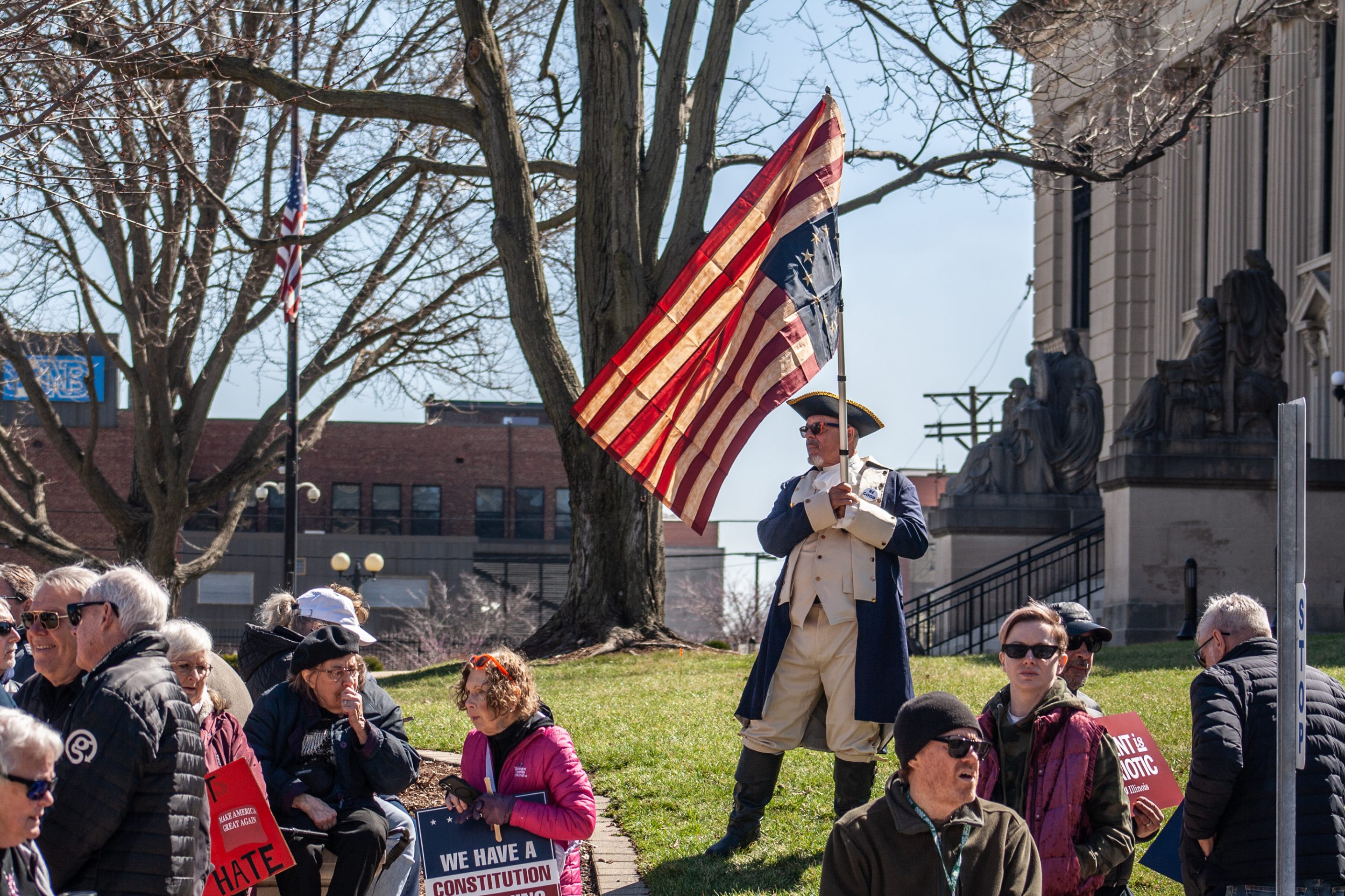 A demonstrator wearing a Revolutionary War uniform holds an upside-down Betsy Ross U.S. flag during a "No Kings" protest outside the Illinois State Capitol in Springfield, Ill., on Saturday, March 28, 2026.