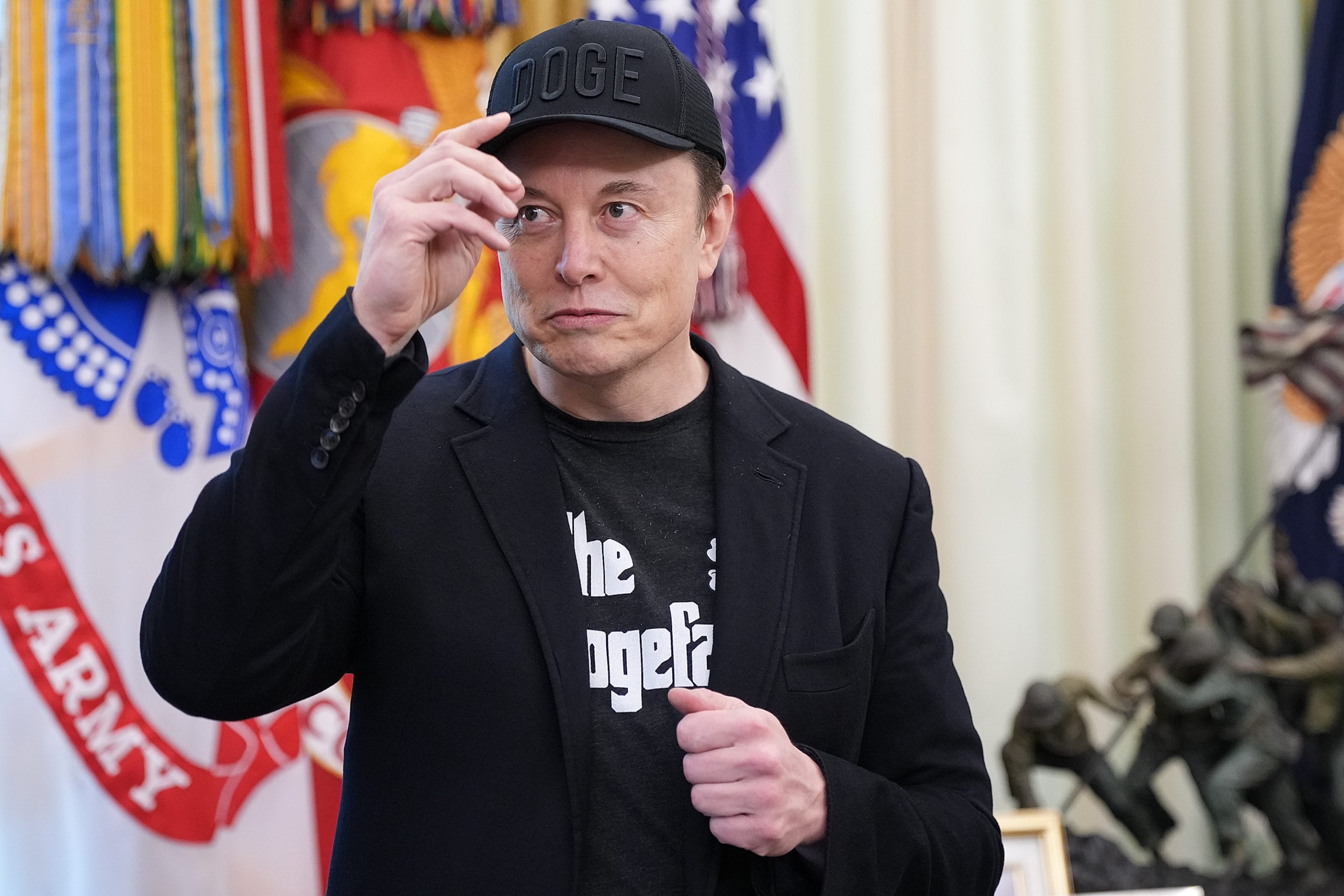 Elon Musk, wearing a DOGE shirt and hat, puts one hand to the brim of the hat as he stands in the Oval Office.