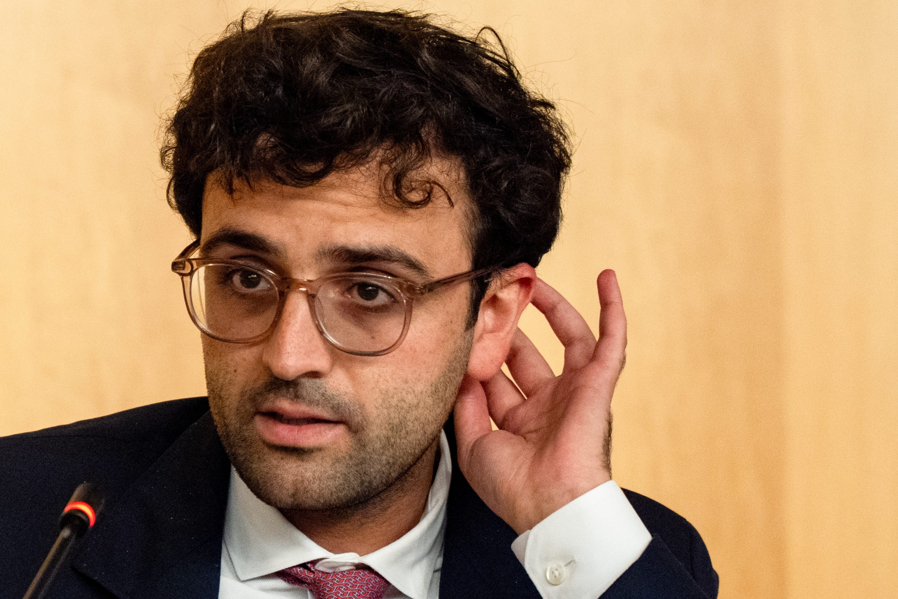 bespectacled man scratches the back of his head during congressional hearing