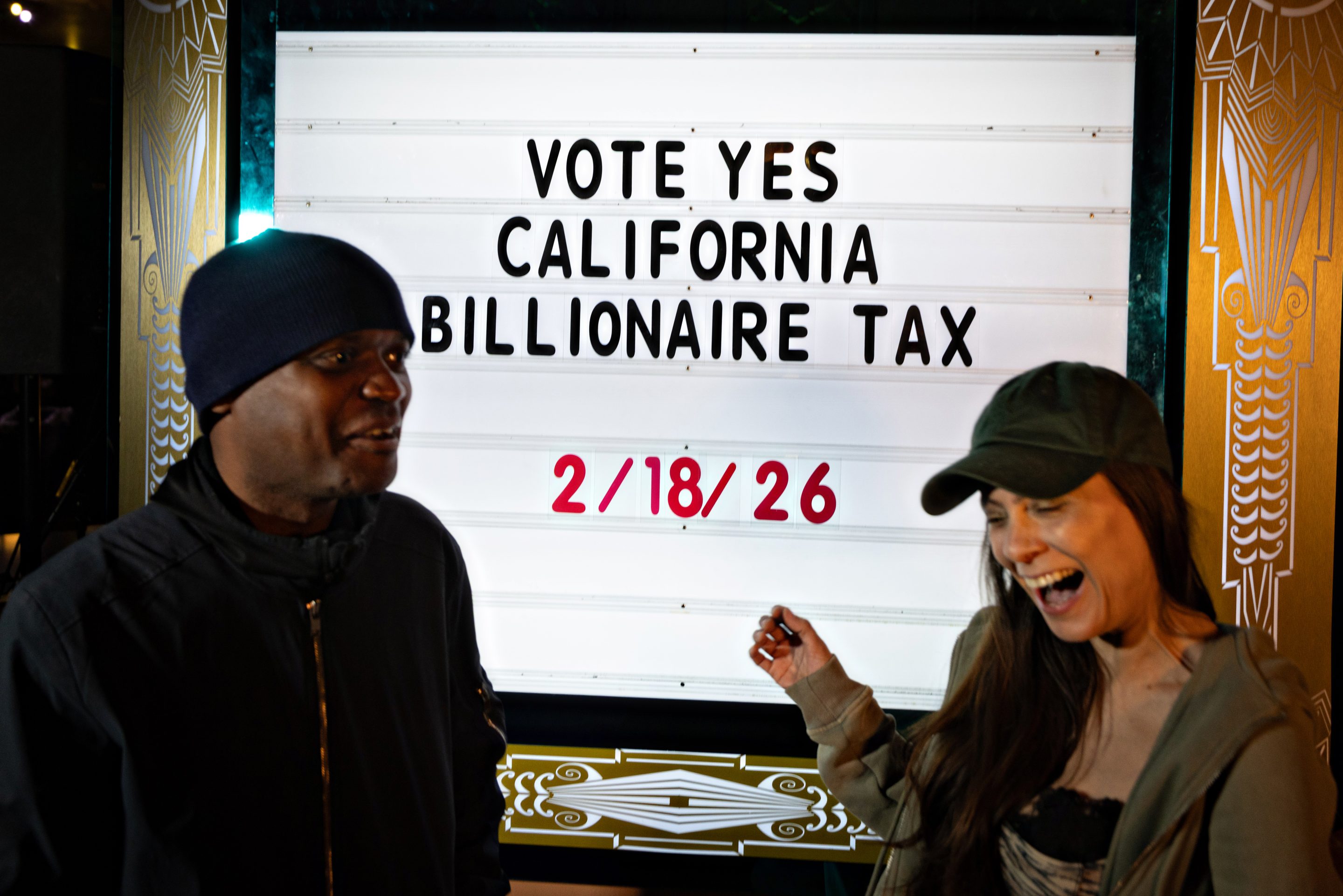 Two people stand in front of a sign urging support for California’s billionaire tax