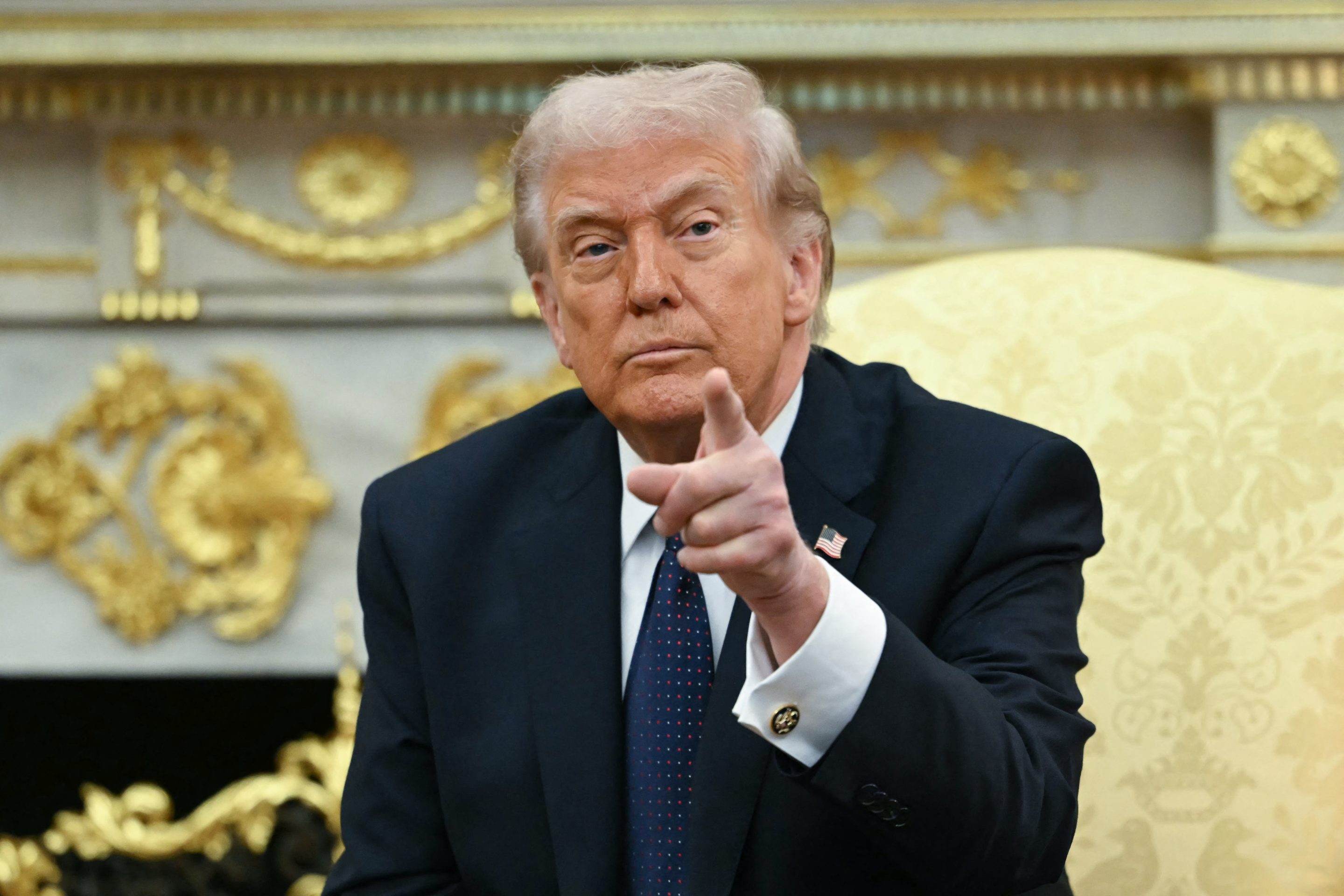 Donald Trump sits in the Oval Office. He looks ahead and points.
