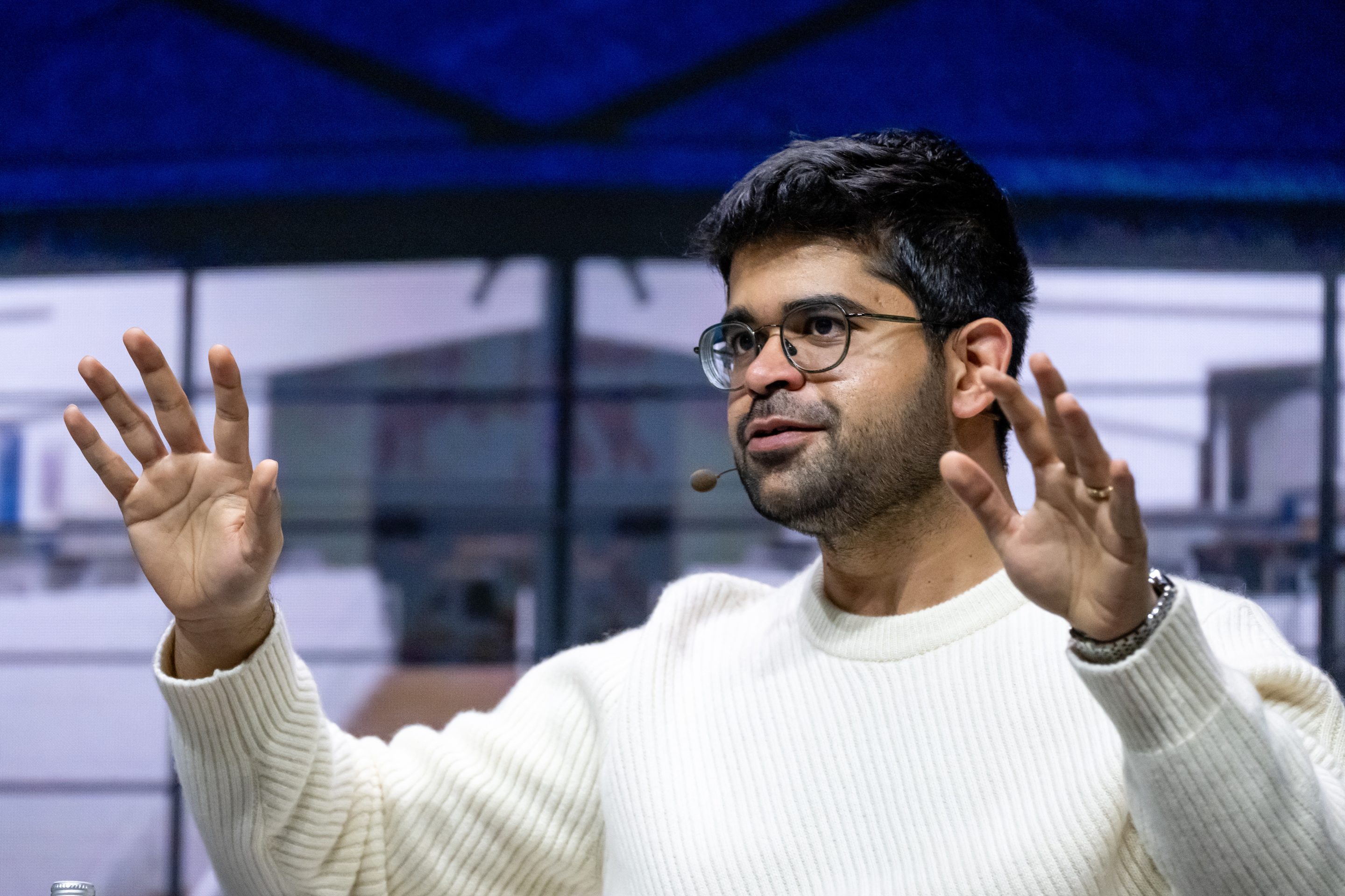 Aravind Srinivas, wearing a white sweater, lifts both of his arms in front of him.