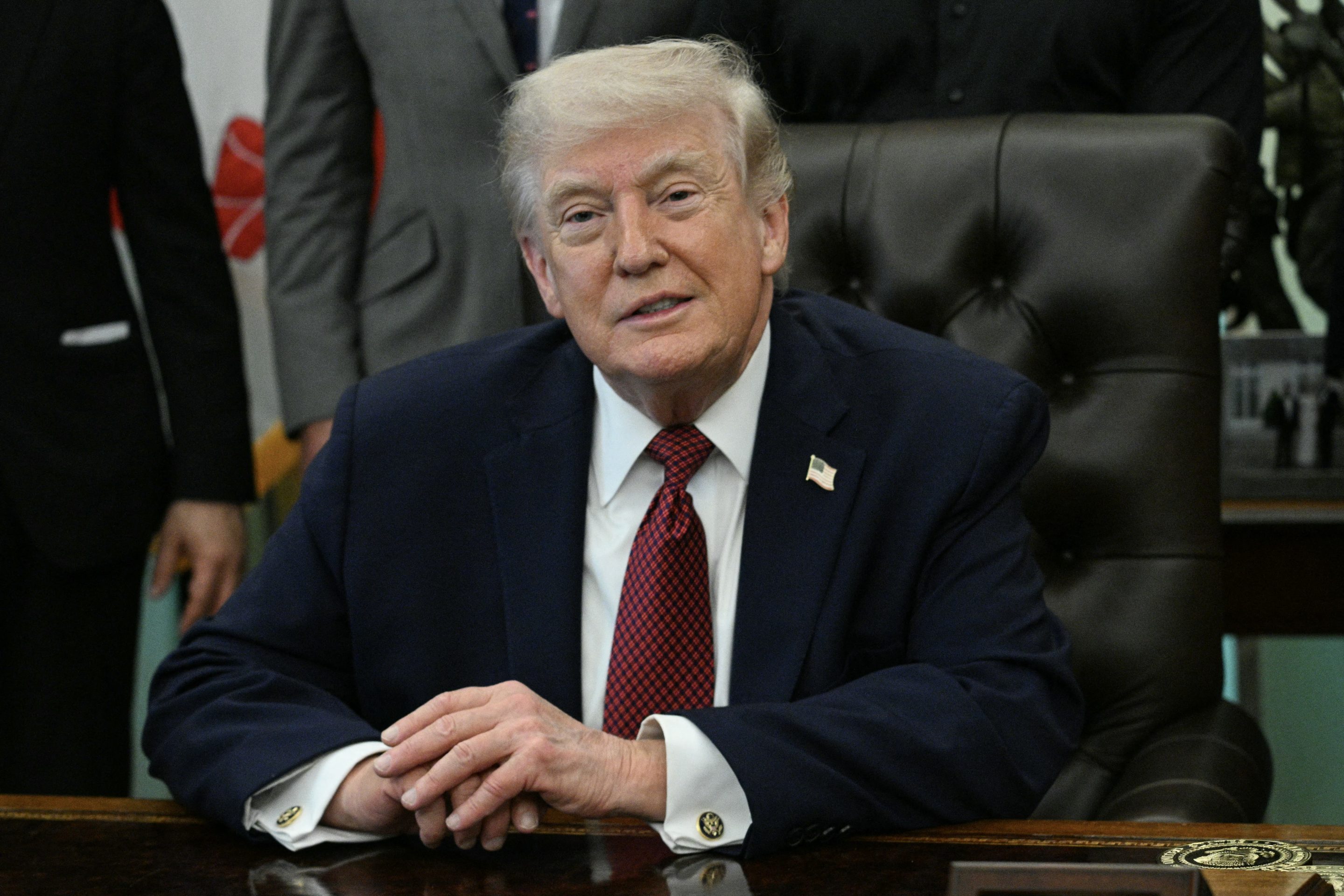 Donald Trump sits at his desk in the Oval Office with his hands folded, smiling.