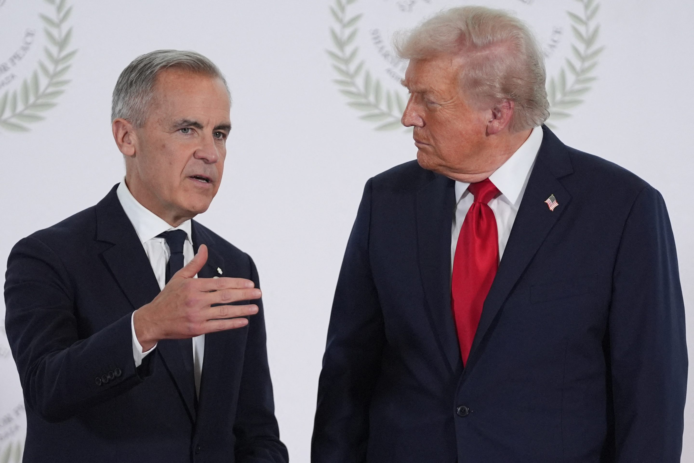 Mark Carney gestures with his hand as he stands next to Donald Trump in front of a white background.