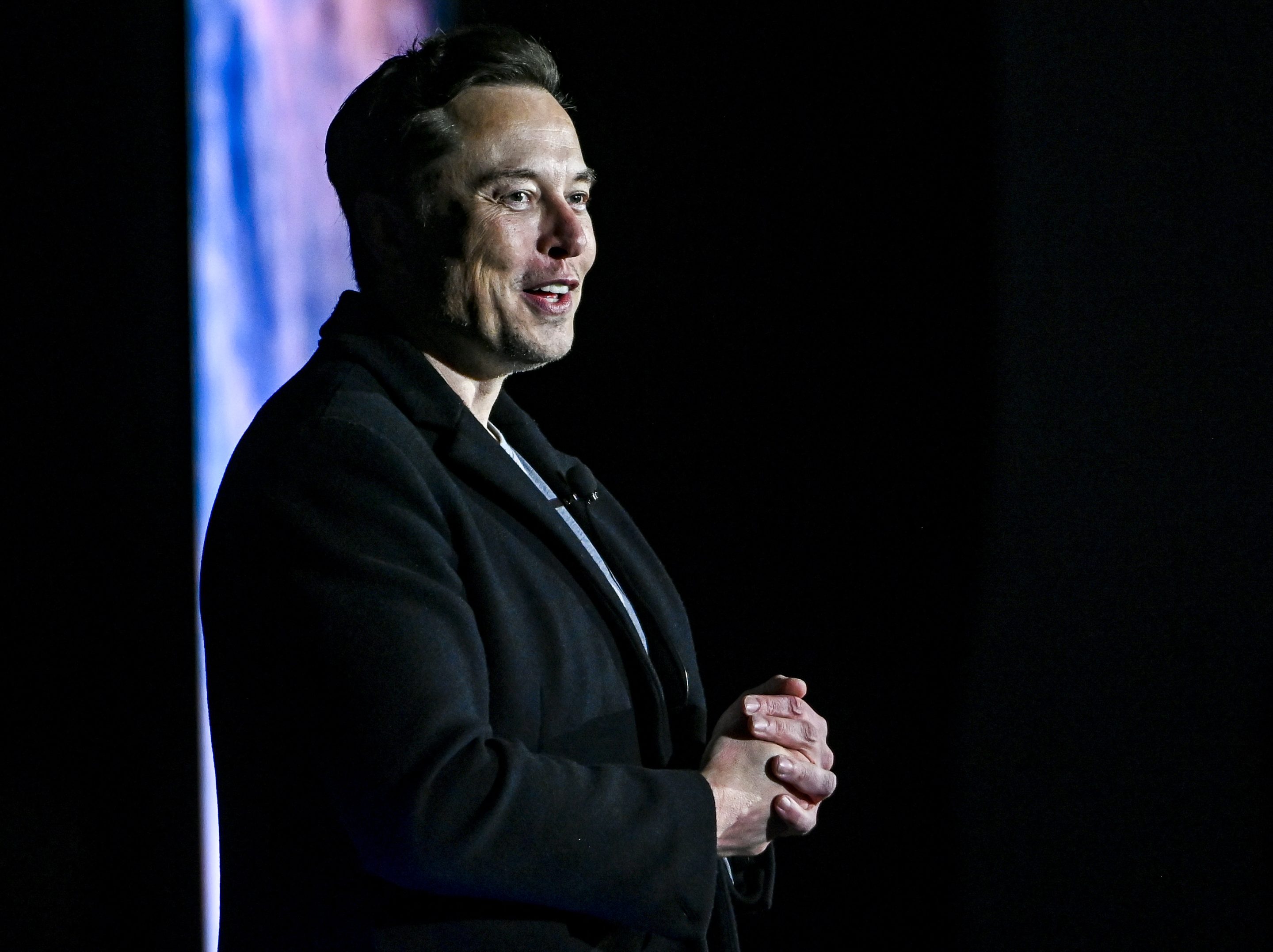 Elon Musk, wearing all black, smiles and looks to the side.