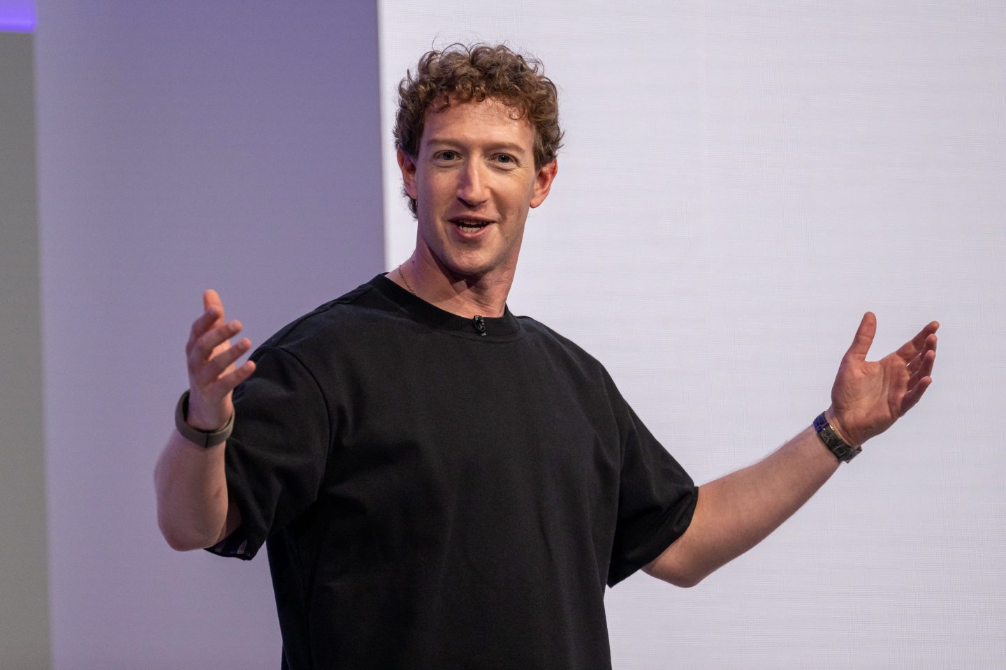 Mark Zuckerberg speaks and holds both hands up while standing in front of a purple background.