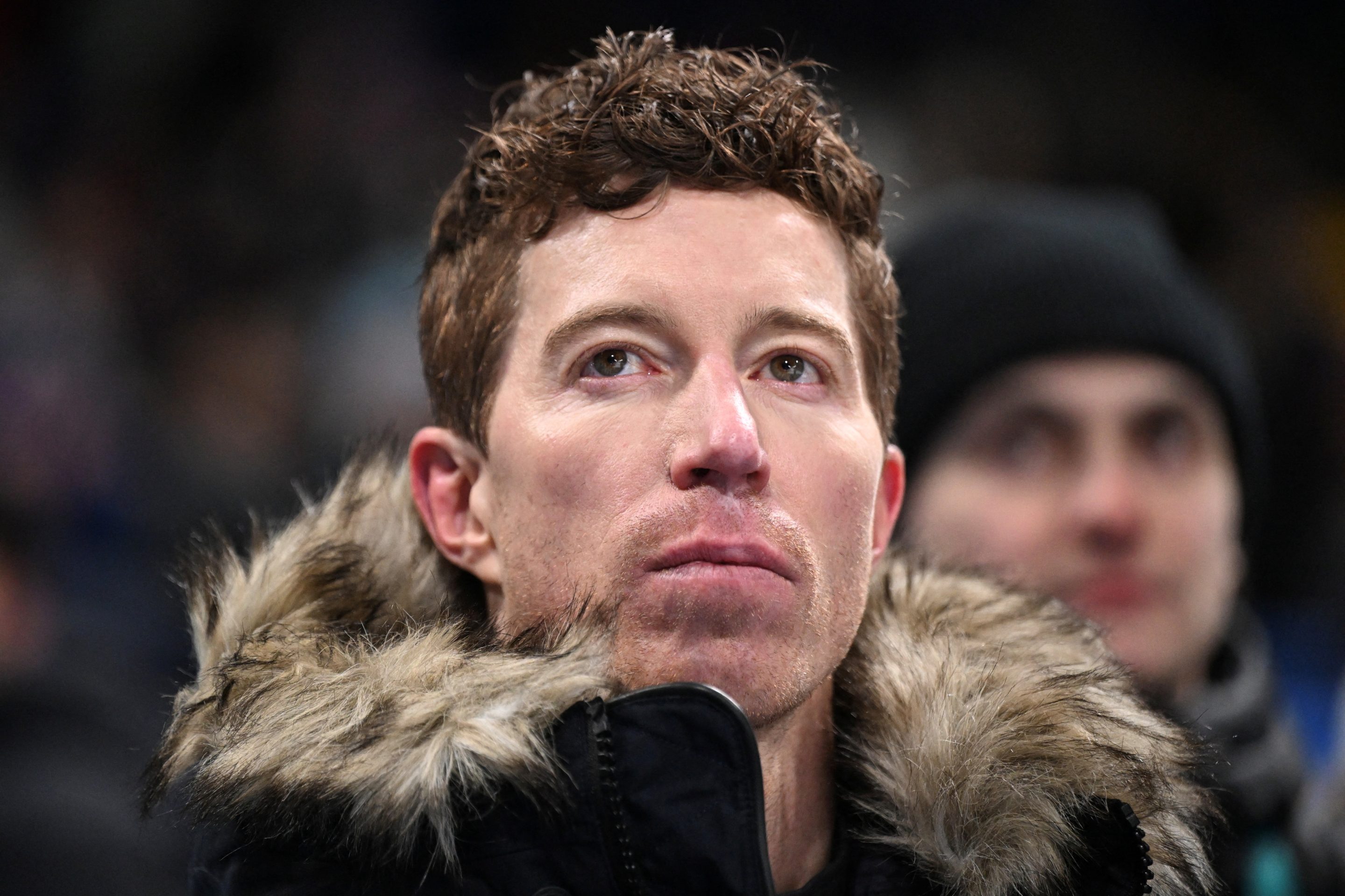 Shaun White, wearing a jacket with a fur-lined hood, looks up.