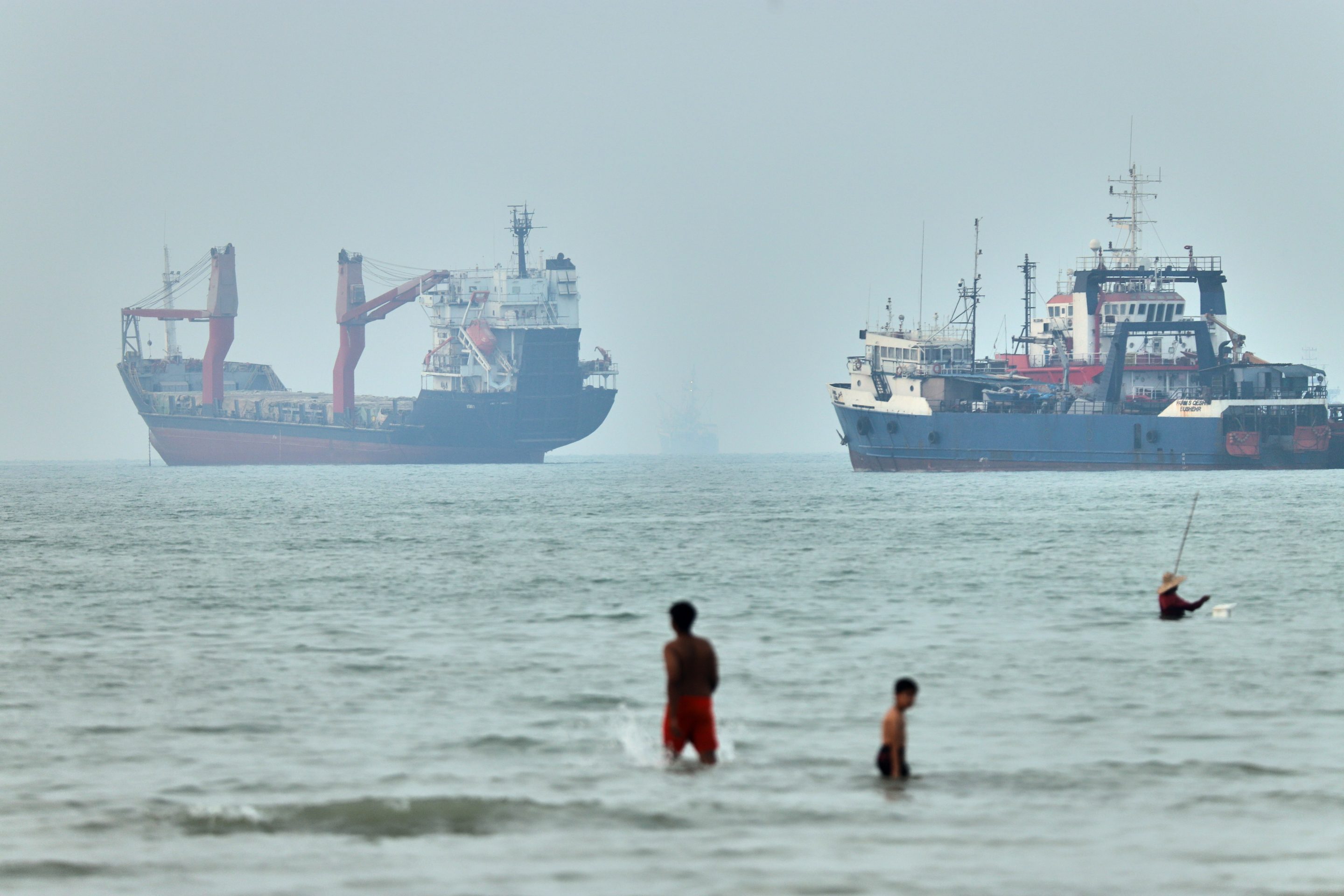 Children wade in the water with cargo ships at anchor in the background and a fisherman nearby, in the Strait of Hormuz off Bandar Abbas, Iran, Tuesday, June 30, 2026.